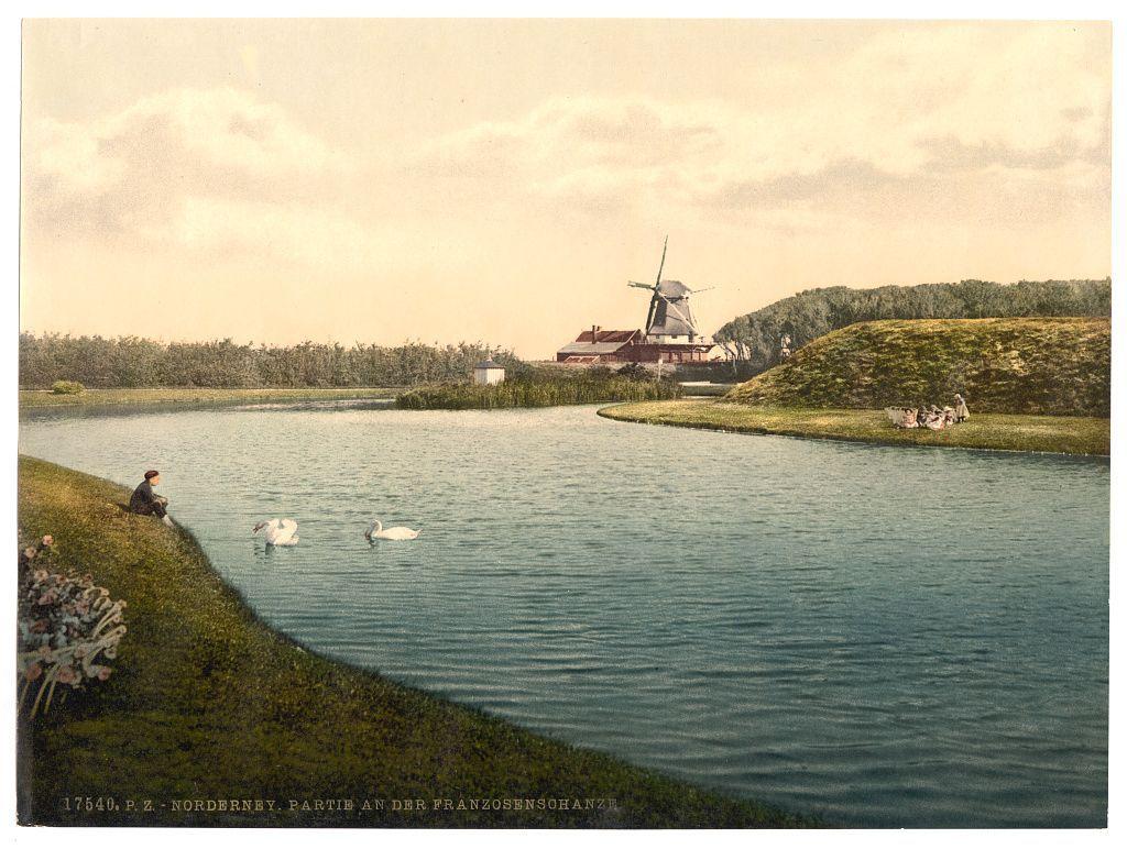 The image depicts a tranquil rural landscape with a calm body of water at its center. On the far side, there is a prominent windmill situated among greenery and trees. The scene includes gentle undulations in the terrain on either bank where people are seated or reclining by the edge of the water. Two white swans float gracefully near the shore as one person sits alone observing them with an open book beside him. Another group appears to be interacting leisurely at a distance, perhaps enjoying a picnic. The overall atmosphere is serene and pastoral, suggesting a time when such scenes were more commonplace in daily life before industrialization fully transformed rural landscapes.