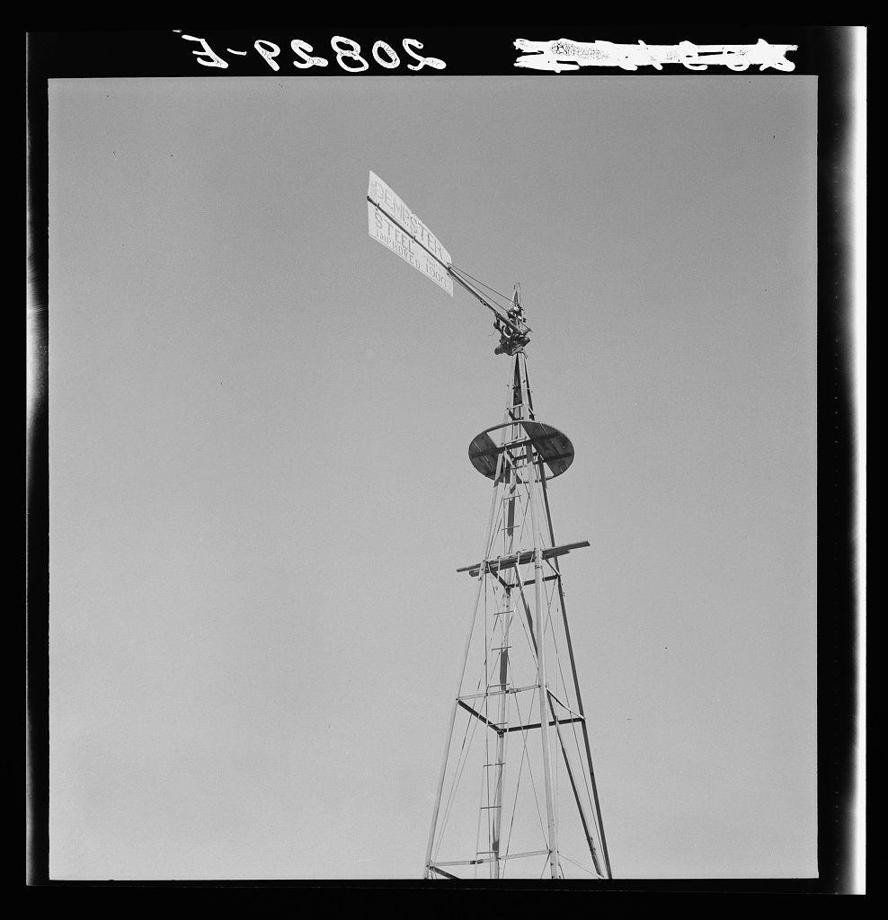 The image captures a tall windmill standing against a clear sky. The structure appears aged and weathered, suggesting it may be an old farm implement or part of a historical site. At the top of the tower is a large metal blade that has likely been used to harness wind power for various purposes such as pumping water or generating electricity on rural farmland.

The photograph itself contains some text elements: "3-P2805" and additional handwritten annotations at the bottom right corner, which might indicate specific references or cataloging details related to Dorothea Lange's photographic archive. The black-and-white nature of the photo suggests it is an older image from a historical collection.

The mention of 'abandoned farm in Columbia Basin' along with Grant County's location and proximity to Quincy likely places this windmill within a rural, agricultural setting in Washington State, USA.