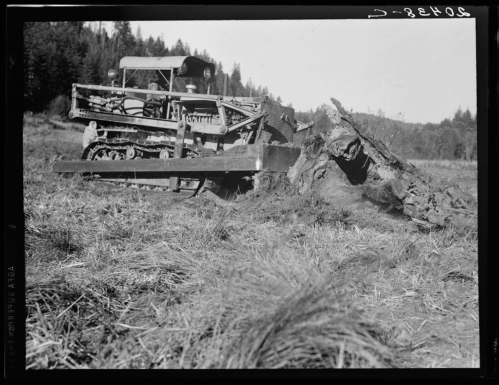 The image is a black-and-white photograph depicting an outdoor scene from what appears to be the early 20th century, judging by the style of equipment and clothing. A large bulldozer with multiple tracks on its wheels dominates the foreground, actively engaged in moving or cutting through dense grasses. Attached to it is a metal structure that seems capable of handling heavy loads.

In the background, a person wearing dark-toned overalls sits atop the vehicle, operating controls for maneuvering and possibly navigating the machine's functions. The landscape suggests rural farmland with no visible buildings except for what could be part of an unseen farmstead or shed in the distant trees on the right-hand side.

The image has text overlay at the top that reads "D-28035," likely a catalog number, and another line across the bottom which is partially obscured but suggests further information about the photo such as date range. The overall impression is one of agricultural labor or land-clearing in progress with mechanized efficiency.