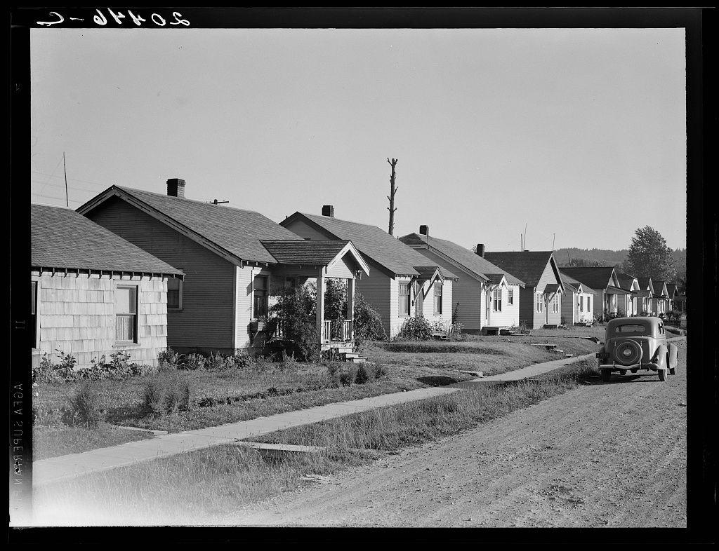 The image is a black and white photograph of a street lined with single-family homes. The houses appear to be modest, primarily one or two-story structures with pitched roofs and varying siding materials including shingles and clapboards. Some houses feature small porches in front, supported by columns. A dirt road runs through the center of the image, curving slightly as it moves out from a viewpoint on the street level.

On this dirt road, an older model car is parked or driving away towards the distance. The surrounding area shows sparse grass and some vegetation between the homes, with patches of bare ground indicating either neglect or limited maintenance. There are no visible people in the image, which adds to its historical ambiance.

The photograph's title "Vancouver Subs" suggests that it was taken in Vancouver, Washington, specifically within Cowlitz County near Longview. The mention of this particular type of housing indicates these homes were constructed by private interests for workers at mills or similar industries during a time when such accommodations would have been necessary to attract labor.

Given the historical context and architectural style, this image likely represents mid-20th-century living conditions in an industrial town setting. It offers insight into working-class housing from that era, with references pointing towards specific locations wher [...]