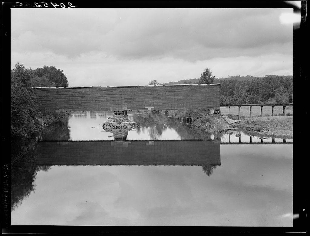 The image depicts a monochromatic view of an old-fashioned covered bridge situated over calm waters. The reflection in the water creates a mirror effect, doubling the visual impact and providing symmetry to the composition. In the background, there's a large brick structure with multiple levels, possibly part of another infrastructure like a dam or lock system for boats. To the right side, we can observe what appears to be wooden railings, potentially forming an observation deck or pathway parallel to the bridge.

The sky is overcast, and clouds dominate much of the upper half of the picture, hinting at possible cool temperatures common in this environment. The overall setting suggests a historical significance, possibly linked with local culture as indicated by references such as Western Washington Thurston County covered bridge on Chehalis River characteristic.