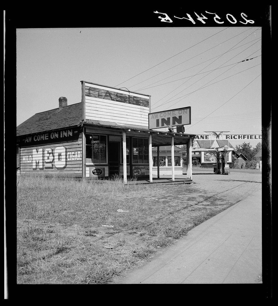 The image depicts a black and white photograph of an old roadside inn, "Welcome Inn," with signage indicating it offers lodging. The establishment has a classic design, featuring wooden paneling on the upper half of its facade and a prominent sign displaying both text and graphic elements including what appears to be a depiction or stylized letter representing 'INN.' To the right stands another structure labeled "Jane Richfield Motel," suggesting accommodation options for travelers in this area. The setting is rural, with an unpaved road stretching from the viewer's perspective into the distance, bordered by grass and weeds indicative of infrequent maintenance. A few sparse individuals can be seen near what seems to be a trash receptacle or similar object on the roadside.

The photograph has been annotated with text in handwriting at the top right corner which may signify "24-hour," potentially referring to one or both establishments being open around-the-clock, although its connection is speculative without additional context. The overall atmosphere of the scene suggests it was captured during a time when such establishments were more common along U.S. highways for travelers making stops on their journeys.

This specific photograph has been associated with Washington State's Lewis County and Centralia region through online sources like https://images.loener.nl/DorotheaLa [...]