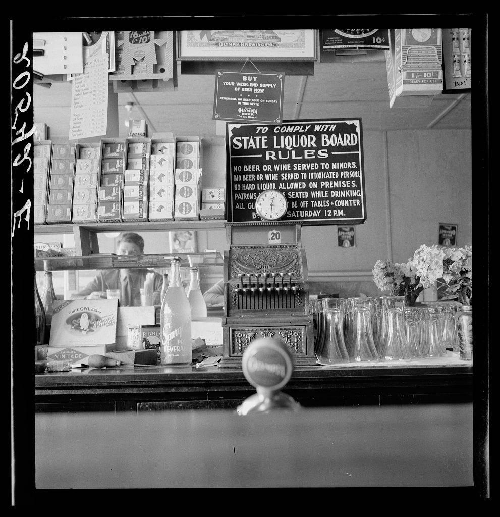 The image depicts an interior scene of a bar or small restaurant, captured in black and white. At the center is a classic-style cash register with dials indicating price levels such as "1," "5," "10," etc., showing that it's set to 20 cents on one dial. Above this counter hangs several boxes presumably containing different types of spirits or tobacco products, labeled for brands like "White Owl Blend" and "Vintage." To the right is a sign from the State Liquor Board outlining regulations about alcohol service; notable rules include not serving beer or wine to minors, no hard liquor on premises after 12 P.M. Saturdays, tables must be off by this time too, among others.

In front of these items are an array of empty glasses and bottles, suggesting a busy establishment. Behind the cash register is a person partially visible, likely attending bar duties. The setting appears dated given its monochromatic quality and style of products displayed.