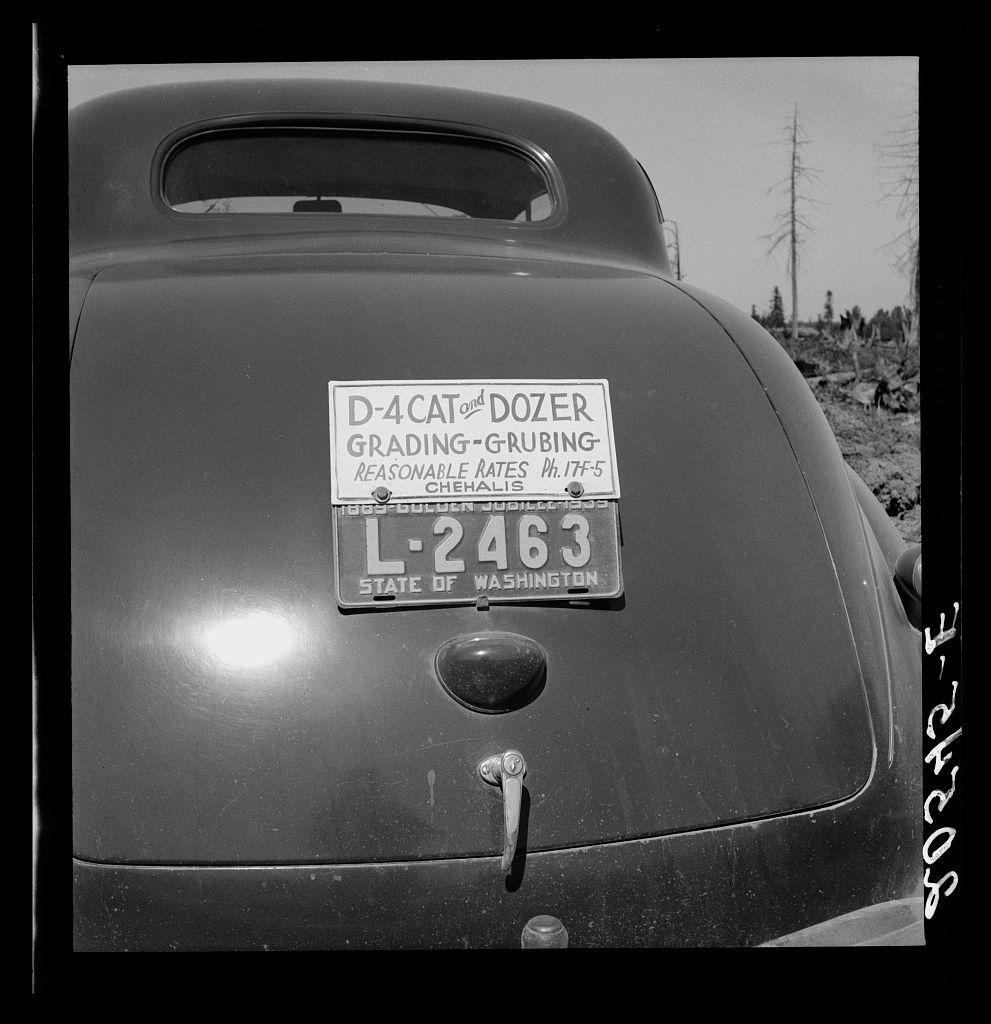 The image depicts a vintage car with several notable features. On the back of this automobile, there is a license plate that reads "D-4 CAT" and below it in larger font, "DOZER," indicating its use for construction purposes such as grading or grubbing roads. The vehicle also has an additional label on the rear window: "State of Washington," suggesting registration within Washington state.

The car's body is a dark color with visible wear marks and minor damage like scratches and dents, which imply it may be quite old and possibly used in tough conditions associated with construction work. A prominent emblem or logo resembling a bulldozer occupies the lower right side on the back of the vehicle, reinforcing its identity as a construction equipment.

In terms of composition, this photograph appears to have been taken outdoors during daylight hours given the brightness surrounding the car's edges and shadows cast by objects around it. The black-and-white tone of the image adds an element of historical context or artistic effect, possibly suggesting that the photo was captured in earlier times when color photography might not be prevalent.

The background is minimal but includes what looks like utility poles with electrical lines running above them against a clear sky, which suggests this vehicle may have been parked along rural roads. No people are visible within the frame o [...]