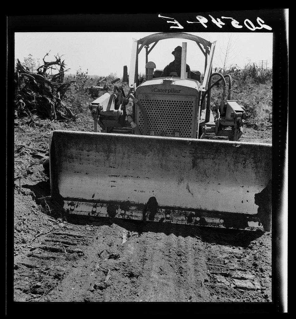 The image captures a scene in black and white, showing an individual operating a large Caterpillar bulldozer. The vehicle is equipped with a grader-type blade at the front, which is used for leveling or smoothing out soil and dirt during construction projects.

In the background, there are remnants of uprooted trees and shrubbery, indicating that this area may have been cleared in preparation for some form of land development. Some vegetation can still be seen further back behind a fence line. The ground around the bulldozer appears to be compacted or churned up from recent activity.

The person operating the machinery is wearing protective gear, including what looks like ear protection and possibly sunglasses or goggles to shield their eyes. They are seated within the cab of the bulldozer, which has an open design with a roof but no side panels for enclosure.

A handwritten inscription on the top right corner reads "E-P-7608," likely serving as identification or cataloging information associated with this photograph. The general caption suggests that more context about the image can be found by referencing number 41 from additional materials related to Nieman farm and Western Washington, Lewis County near Vader.