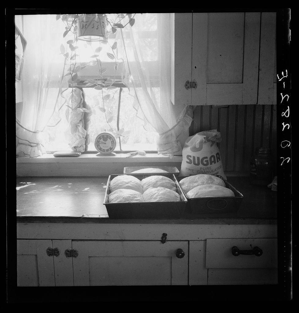 The image captures a vintage-style kitchen scene filled with an assortment of objects that evoke a sense of nostalgia. The primary focus is on the window sill, where various items are neatly arranged in front of what appears to be frosted glass curtains. To the left side, there's a decorative lampshade hanging over a jar and two plates stacked atop one another.

On the right side of the window sill sits an old-fashioned clock surrounded by delicate lace or ribbons that seem to cascade down from above, creating a picturesque display reminiscent of early 20th-century domestic interiors. Adjacent to these items is a sack labeled "SUGAR," signifying its contents and adding to the vintage atmosphere.

In front of this assortment lies an open box containing several round loaves of bread or rolls, placed on what seems like a wooden surface with drawers beneath it. The entire scene exudes warmth and homeliness, suggesting that someone has just been busy baking in preparation for perhaps breakfast or another meal.

The black-and-white color scheme enhances the timeless quality of the photograph, drawing attention to textures such as the rough fabric of the sugar sack, smooth surfaces like polished wood, and softer elements like lace. The overall composition suggests a domestic setting, with an emphasis on simplicity and tradition rather than modernity.