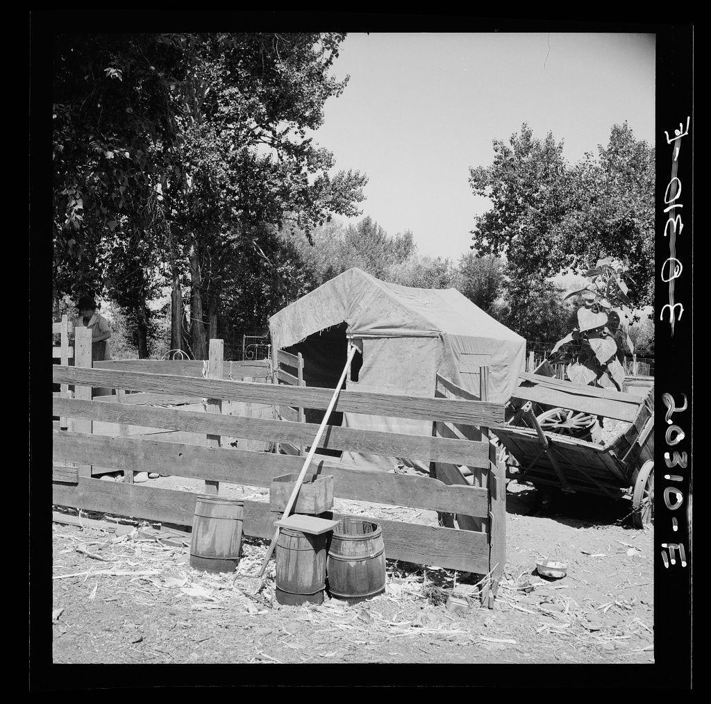 The image is a black and white photograph depicting an outdoor setting with rustic elements. In the foreground, there's a wooden fence constructed from horizontal planks supported by vertical posts. Leaning against this fence are two barrels on their sides; one barrel appears to be overturned. Next to it stands another object that looks like a makeshift stool or container.

A central feature of the image is a tarp-covered structure with an open front, revealing what might be a wooden interior frame inside. The fabric covering seems weathered and partially damaged at the top edge where some sections are frayed or missing entirely, exposing part of the framework underneath.

On the right side of the photo stands another makeshift object that resembles a covered wagon or cart, possibly constructed from planks with metal supports. It's positioned in an uneven manner on what appears to be muddy ground strewn with debris and small branches.

The background includes trees which suggest this setting is located near a wooded area. The sky is visible through the gaps between tree branches but it's not clear whether there are clouds or if it's bright daylight. There's also some form of writing in the upper left corner that reads "3-9-10-E," possibly indicating a date and reference code, hinting at archival documentation related to this scene.

Overall, the image portrays an impressi [...]
