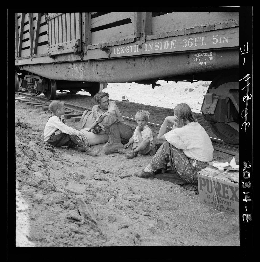 The image depicts a group of people gathered on the ground near a freight train. There are four individuals, likely family members, sitting or squatting around each other in a circle-like formation. One child is seated on what appears to be a rolled-up rug or blanket while another younger individual sits beside an adult who seems engaged with them. Another person leans against something out of the frame with their hands clasped together near their chest.

The setting suggests they are outdoors, as there's sand visible in the foreground and no shelter overhead is apparent. The ground appears to be a mix of soil or dirt and small rocks, indicating it could be an unpaved area alongside train tracks. Behind them, part of a freight car with text on its side can be seen; however, most of the details are not clear enough for precise transcription.

The image captures this scene in black-and-white tones, which gives it a historical or nostalgic feel and highlights contrasts between light and shadow effectively. The composition includes personal items like an open box labeled "PUREX" with partial text suggesting its contents relate to dental care products, hinting at the necessity of such goods for those traveling on trains.

The overall atmosphere evokes a sense of informality as they rest or wait together amidst their travels by freight train in Washington state.