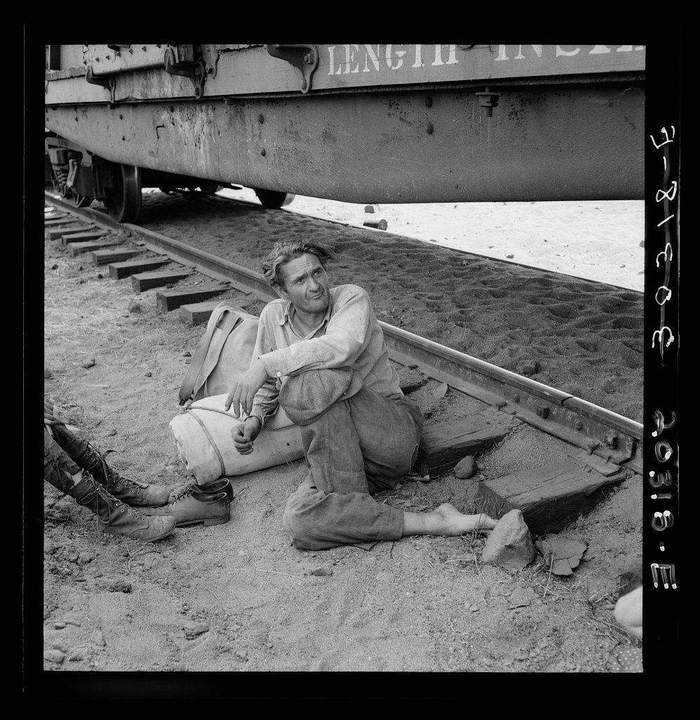 The image is a black and white photograph showing an individual sitting on the ground beside railroad tracks. The person appears to be engaged in some sort of labor, possibly related to railway work or maintenance, as suggested by their posture and attire which includes worn-out clothing and boots typical for manual jobs.

They are seated with one leg bent at the knee and foot resting on a wooden plank that runs parallel to the train track beside them. The other leg is stretched out towards an open railcar labeled "ENGINE 107," indicating it may be part of a freight or cargo transportation system.

The individual has disheveled hair, possibly unkempt due to work-related activities or fatigue. Their expression seems contemplative or weary as they gaze off into the distance with their head tilted slightly downwards and eyes not directly engaging with the camera lens.

In front of them is what appears to be a rolled-up canvas bag or blanket resting on the ground beside another smaller sack-like object, possibly containing personal belongings or tools related to work. The environment around shows rough terrain typical for railway tracks, with visible sand or dirt scattered across the area and wooden planks used as makeshift paths.

The photograph's overall atmosphere conveys hardship, laborious conditions, and perhaps a transient lifestyle associated with traveling from one p [...]