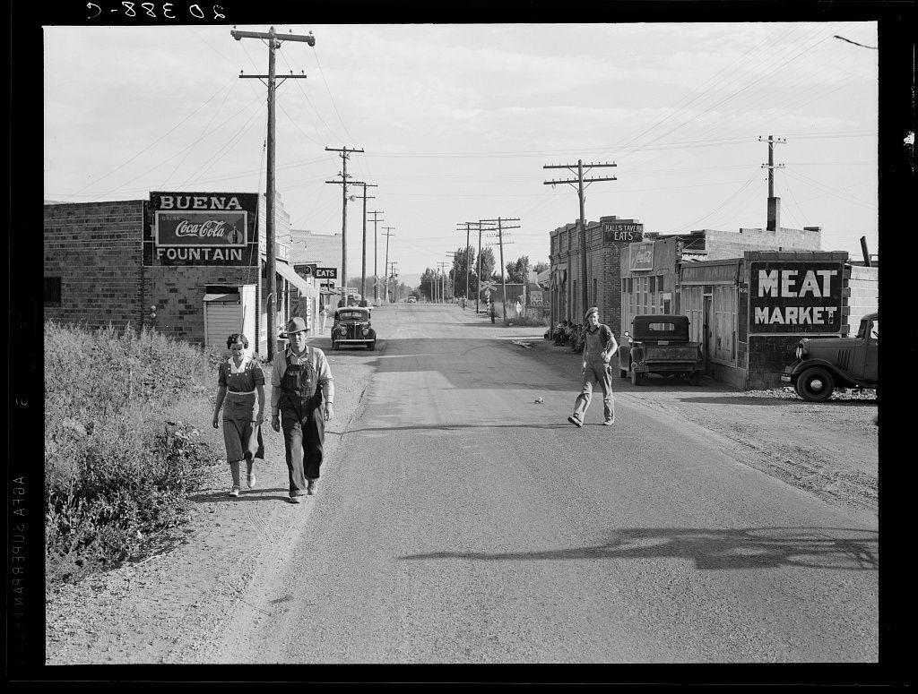 The image captures a moment from what appears to be mid-20th century life in an American small town. It is presented as black and white, adding historical context with its grayscale tones.

On the left side of the street stands a building labeled "BUENA COCA-COLA FOUNTAIN," suggesting it was likely a place where people could get refreshments including Coca-Cola products. The sign indicates that this establishment also served food at 'EATS,' as indicated by another part of the sign which is partially visible.

On the right, there's an old-fashioned truck with "MEAT MARKET" written on its side, indicating it was used for delivering meat to customers or possibly transporting goods within town. Behind it stands a structure that seems to house more businesses: one reads 'HALF-STRAW BAKERY' and another has signage partially hidden behind the man who is walking.

Two people are seen walking down the road; on the left, there's an individual wearing what appears to be work overalls with suspenders, hinting at a farmer or someone working in agriculture. The person seems focused forward as they walk alongside them.

On the right side of the street walks another man who looks like he might have been involved in some manual labor; his attire is more casual than that of the first individual and includes what appears to be overalls with rolled-up sleeves, possibly indicating a hands-on  [...]