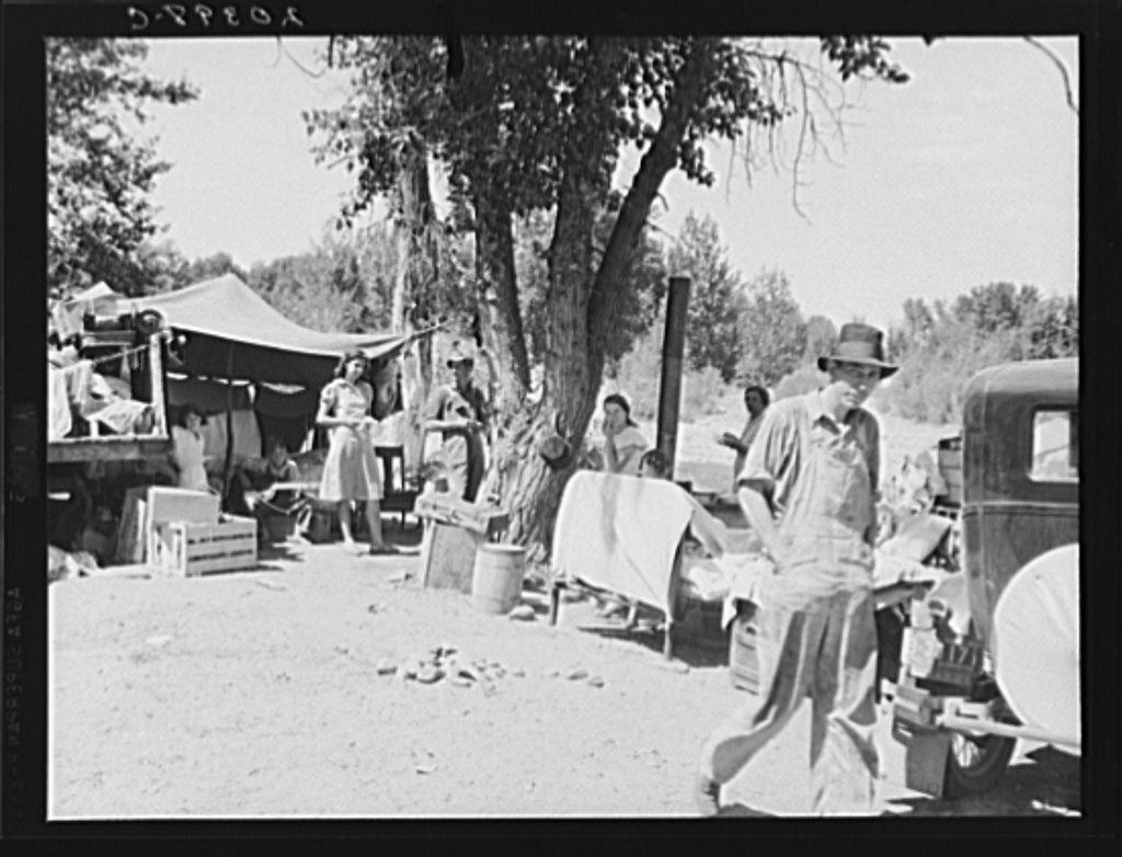 The image is a black and white photograph depicting an outdoor scene with multiple individuals, likely from the early to mid-20th century judging by clothing styles. A man in overalls stands prominently at the forefront wearing a hat, suggesting he might be of importance or simply dressed for protection against sun exposure.

In the background, several people are engaged in various activities near tents and makeshift structures that suggest a temporary settlement possibly related to migrant work camps such as "Ramblers Park" mentioned in the caption. The environment appears rural with trees providing shade over some areas while others remain exposed under direct sunlight. A vintage car is also visible on the right side of the image, reinforcing the historical context.

The overall atmosphere conveys hardship and resilience among individuals within a nomadic or transient community setting during times when labor migration was more common due to economic factors such as agriculture seasons or industrial developments requiring seasonal workers.