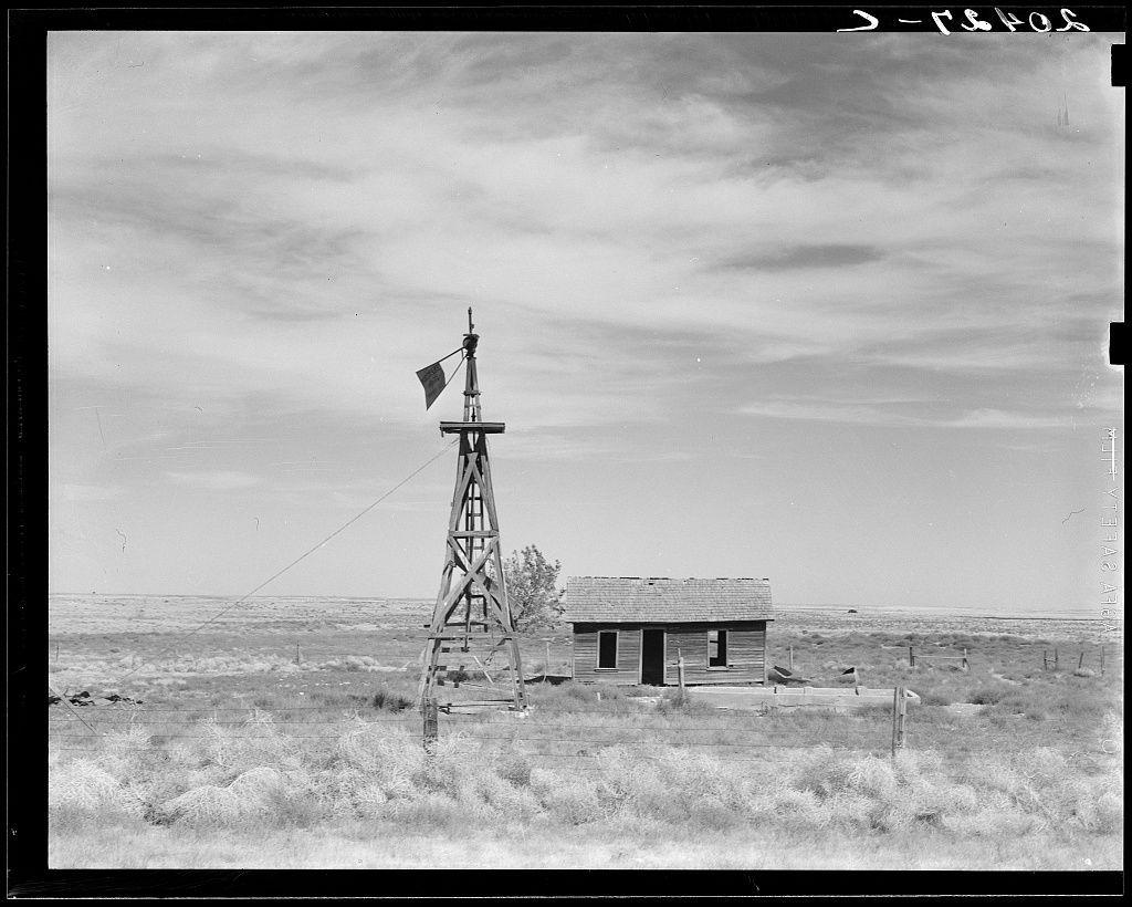 The image shows a desolate rural scene with an old windmill and weather vane situated in the foreground. The structure appears to be made of wooden planks, standing alongside what looks like a small shack or outbuilding with boarded-up windows, suggesting abandonment or disuse. In the background is an expansive landscape characterized by dry shrubbery indicative of arid conditions, likely part of a larger desert environment. Above, a cloudy sky stretches across much of the image's upper half, contributing to a sense of openness and isolation. The photograph has been taken in black and white, giving it a timeless or historical quality that enhances its depiction of rural hardship or decay.