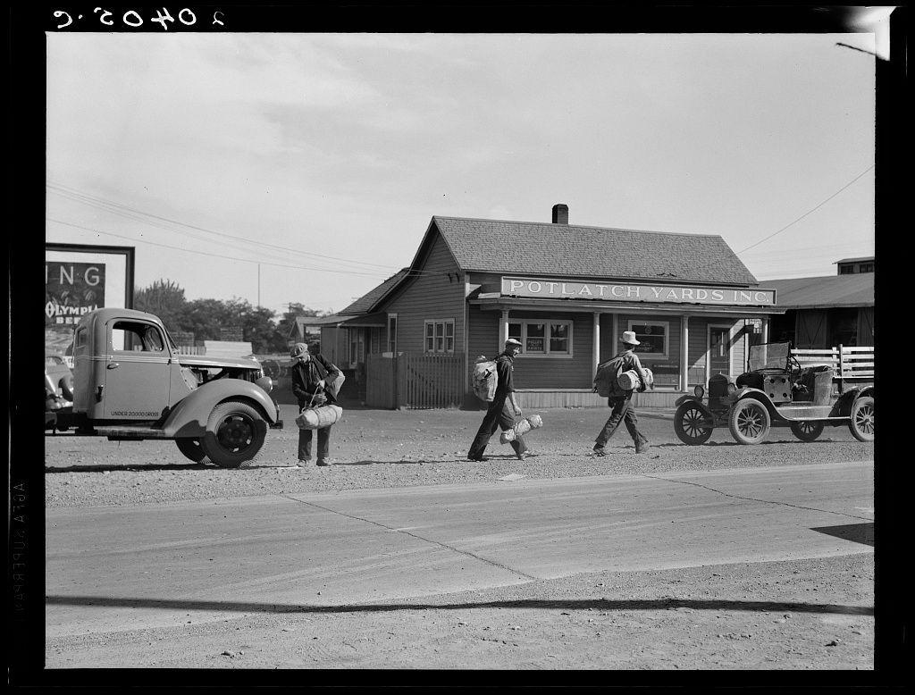 The image is a black and white photograph depicting a street scene from the early to mid-20th century. It shows three individuals walking across a paved road in what appears to be an industrial or commercial area, given the presence of various buildings with signs such as "Potlatch Yards Inc." The people are dressed in casual work attire and appear to carry items that might suggest they are traveling for business purposes.

In the foreground, there is a vintage truck parked on the side of the road. Behind it stands an old-fashioned building labeled "Olympia," possibly indicating a store or gas station from that era. Another older vehicle, likely an antique car with spoked wheels and early design features, is also visible in the background.

The overall atmosphere suggests a snapshot of daily life during a bygone time, capturing elements typical of rural America at the time when railroads were vital for transportation and commerce.