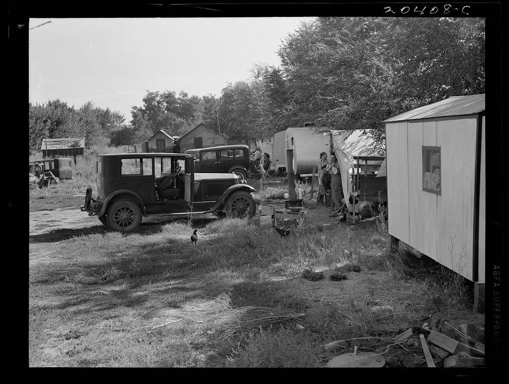 The black and white photograph depicts a rural setting with several old-fashioned cars parked on the grass. There are trailers or small sheds in the background, along with some trees lining the area. People can be seen standing around near the vehicles, while another individual is walking away from them. A chicken stands prominently in front of one car, adding life to the otherwise still environment. The image captures a moment that seems like it's taken back in time, reflecting on a simpler way of living and perhaps providing insight into migratory workers' lives during different seasons or periods.