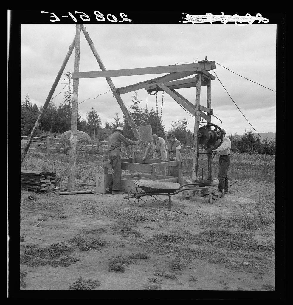 The image depicts a historical scene of individuals engaged in manual labor, using mechanical equipment to drill or extract water from the ground. It is a black and white photograph with monochromatic tones that convey a sense of timelessness. The setting appears rural, possibly on farmland given the presence of wooden fencing and sparse vegetation indicative of an outdoor environment.

Several people are visible; one person seems to be operating a large mechanical device mounted on stilts or a frame, which could be part of a drilling rig used for well construction. Others appear focused on assisting with various tasks related to this operation. There is also equipment such as wooden planks and metal tools that suggest manual labor involved in the process.

The landscape features an open field with scattered vegetation, hinting at rural or agricultural surroundings. The sky seems overcast, suggesting a cloudy day which adds to the historical ambiance of the scene.

In terms of context provided by the caption, this photograph is associated with non-commercial experiments on farming loans facilitated by the Farm Security Administration (FSA) in Michigan Hill, Thurston County, western Washington State during what could be around late March or early April 1930. The image and its historical significance likely provide insights into rural practices of well construction for agri [...]
