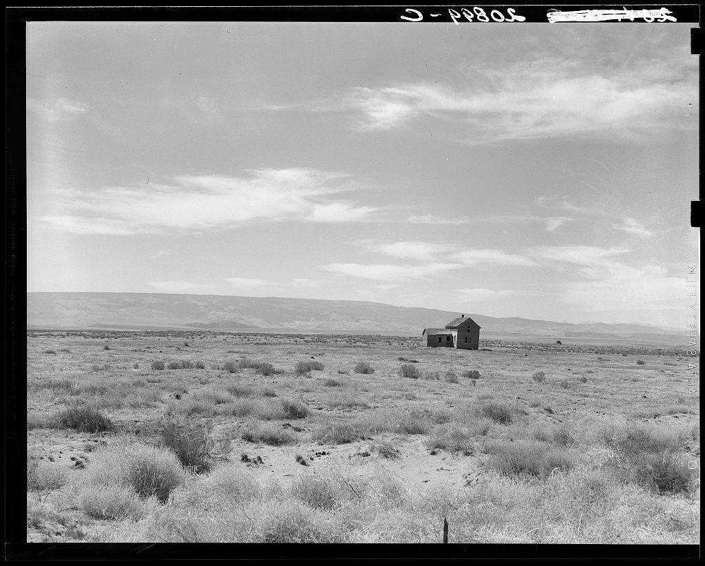 The image is a black and white photograph depicting an abandoned dry land farm situated in the Columbia Basin, South of Quincy, Grant County, Washington. The scene captures a large expanse of arid landscape with sparse vegetation consisting mainly of low-lying shrubs or grasses spread across the terrain.

In the center of this expansive field stands an isolated building that appears to be derelict and unused for its original purpose. It is relatively small compared to the vastness around it, emphasizing a sense of isolation in the rural setting. The sky above spans out into the distance with soft clouds scattered about, suggesting a calm but desolate atmosphere.

The absence of human activity or modern equipment suggests that this area has been left untouched for some time. This could be indicative of economic challenges faced by farmers in such regions during times when agriculture became less profitable due to factors like overuse of arable land and declining water availability from the basin's rivers, which is a common issue in many rural American communities.

The photograph evokes feelings of nostalgia or perhaps a reflection on past agricultural practices and settlements that have since been overtaken by nature. The simplicity of the scene draws attention to themes such as abandonment, resilience against droughts, farming economies' fragility, land use challenges fa [...]