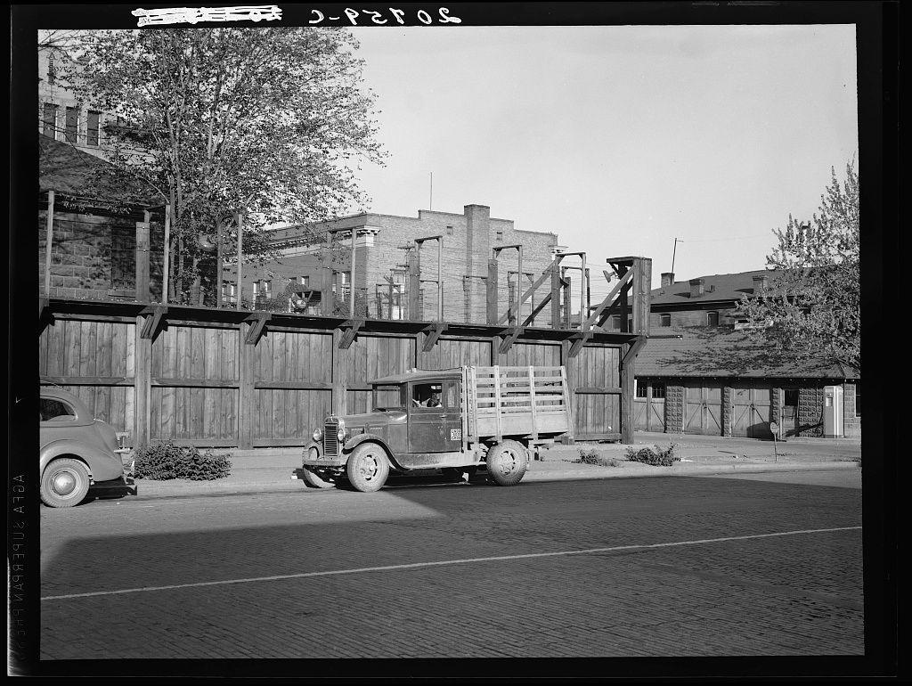 The image is a black and white photograph depicting an urban street scene. In the foreground, there's a paved road with two lanes marked by parallel lines. On the left side of the road, we see part of another vehicle—a vintage car from what appears to be mid-20th century—parked on the curb. The right lane is occupied by an older model truck parked in front of a large wooden fence.

The fence itself seems old and weathered, with several vertical boards supported by concrete or stone blocks at intervals along its length. It's part of what looks like industrial buildings behind it, one of which has tall chimneys indicating possible factory use. The architecture suggests an older urban area possibly from the early to mid-20th century.

In front of these structures is a large wooden platform with railings and a walkway leading up its side. It resembles some sort of loading dock or industrial equipment used for handling materials, given its sturdy construction and elevated design. No people are visible in this photograph; there's no indication of activity other than the parked vehicles.

This scene could be interpreted as part of an urban landscape with remnants from a bygone era—perhaps indicative of post-industrial decline or preservation efforts to maintain historical architecture amidst modernity.

The image is labeled "Untitled photo, possibly related to: Washington, Yakim [...]