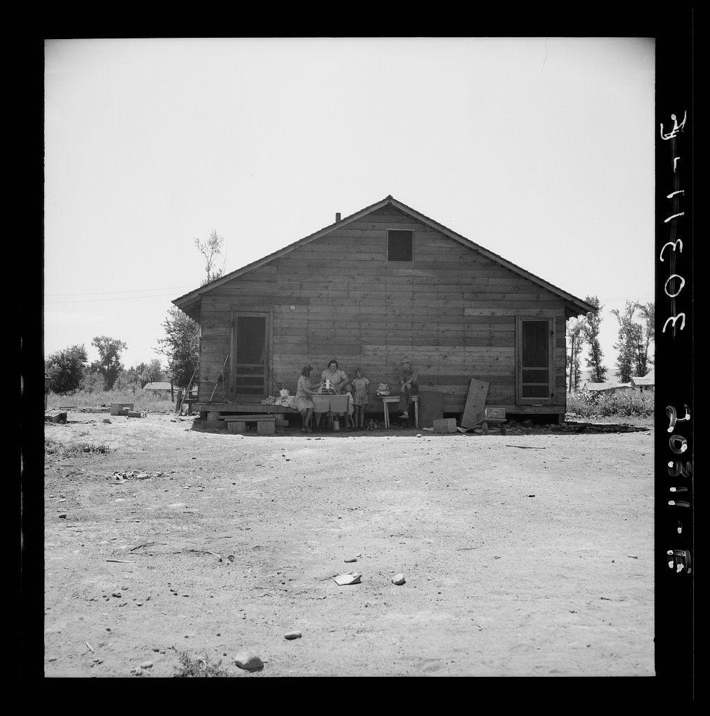 The image depicts an old, rustic wooden cabin in what appears to be a rural setting. The structure has a prominent gabled roof and is constructed from horizontal wood planks with visible weathering and aging signs such as paint peeling off the exterior walls. In front of this house, there's a family seated on a bench that spans across part of its porch area.

The immediate surroundings are barren, with sparse vegetation including small trees and bushes in what seems to be dry soil scattered with rocks. The ground is mostly dirt or sand, indicating arid conditions possibly typical for the Pacific Northwest region during certain times of year due to droughts common there.

There's a sense of poverty conveyed by the lack of visible amenities such as outdoor furniture beyond the bench, and various household items like buckets are placed haphazardly around. The family appears to be engaging in casual activities; some individuals seem focused on tasks or conversation while others appear more relaxed.

The photograph is black-and-white with high contrast, suggesting it was likely taken during a period when color photography was not widely used for documentary work, potentially mid-20th century based on the style of clothing and general quality of life depicted. There's an informative note in white text overlaid at the edge of the image which might provide context or backstory ab [...]