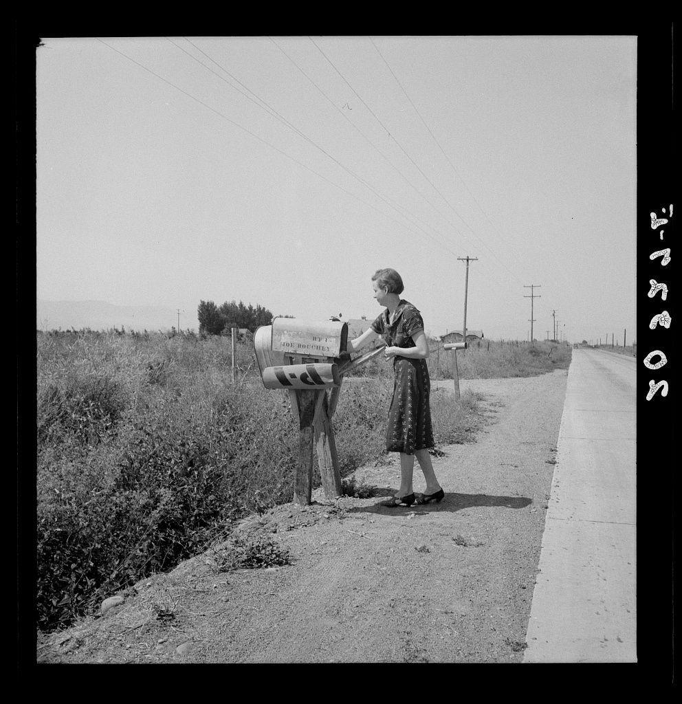 A black and white photograph depicts a scene from what appears to be the early mid-20th century. It shows an adult woman, likely in her 40s or older, standing beside a mailbox on a rural road that cuts through open fields with patches of vegetation. The woman is dressed in a knee-length floral dress paired with dark shoes and holds something in her right hand which seems to be either mail she has retrieved from the box or perhaps an item used for handling such tasks like a pair of gloves.

The mailbox, mounted on a wooden post, bears the name "Joe Bouchey" along its side. It is situated by the roadside where there are visible telephone poles and lines stretching into the distance, suggesting rural connectivity but no immediate signs of residential development close to hand. The sky above is clear with no clouds in sight.

The photograph captures a moment that may be part of daily life for this individual who seems to be attending to her mailbox at what could potentially indicate morning time given it's described as 'the morning mail'. There are elements such as the rural environment, style of clothing and hairstyle which together give an impressionistic idea about the socio-economic status or region where she lives. The photo is credited under "Untitled" with a mention that possibly relates to Mrs. Bouchey getting her daily mail in Washington's Yakima Valley near Toppenis [...]