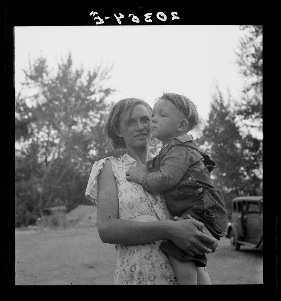 The image is in black and white. It depicts a woman holding a young child, who appears to be looking at something off-camera with curiosity or interest. The adult has short hair that falls slightly past her ears and wears a sleeveless dress adorned with circular patterns. She holds the child close to her chest, providing support as they stand outside during what looks like daytime.

The background shows an open area with trees scattered around, suggesting a rural setting. Behind them is another individual partially visible in profile, wearing overalls. Further back, there's a vintage automobile and other indistinct figures or objects, contributing to the sense of being outdoors possibly near a community gathering spot or on a road.

The photo has an inscription at the top that reads "3-ye2os." The bottom right corner contains additional text: it says "Untitled" followed by some characters and numbers. This could be part of a larger caption explaining more about the context, but only these words are visible in this particular cropped section of the image.

Overall, the photograph captures an intimate moment between two individuals amidst what seems to be everyday life during a specific time period suggested by its monochromatic nature and style of clothing worn.