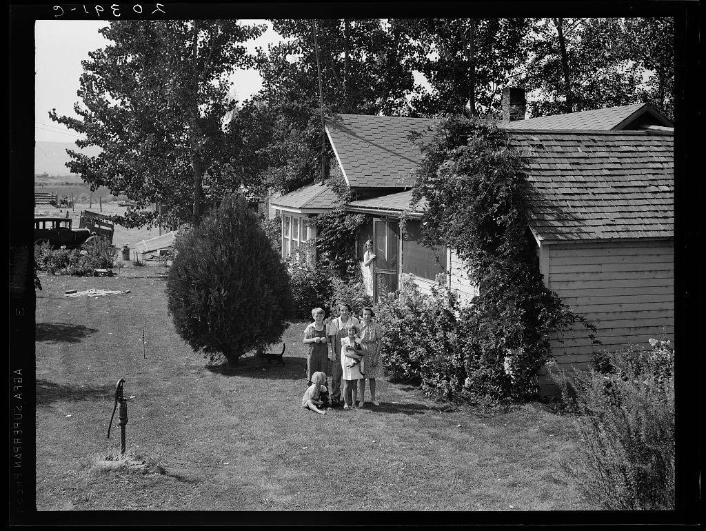 The image depicts a black-and-white photograph of an old house with the label "Farm Security Administration" at the top, indicating it was part of a project to document rural life and agricultural communities in America during that time. The structure is two-story wooden construction with shingle roofing, surrounded by large trees and abundant greenery.

A family consisting of several adults and children stands together outside on what appears to be their property or yard. One adult holds an infant while another woman sits beside them; one child crouches near a small garden fork in the foreground. The attire suggests it's from earlier decades, possibly mid-20th century.

In the background, additional houses are visible along with open fields and more vegetation, implying rural surroundings. A car is parked farther away on what appears to be an unpaved road or driveway leading towards other structures within this community setting.