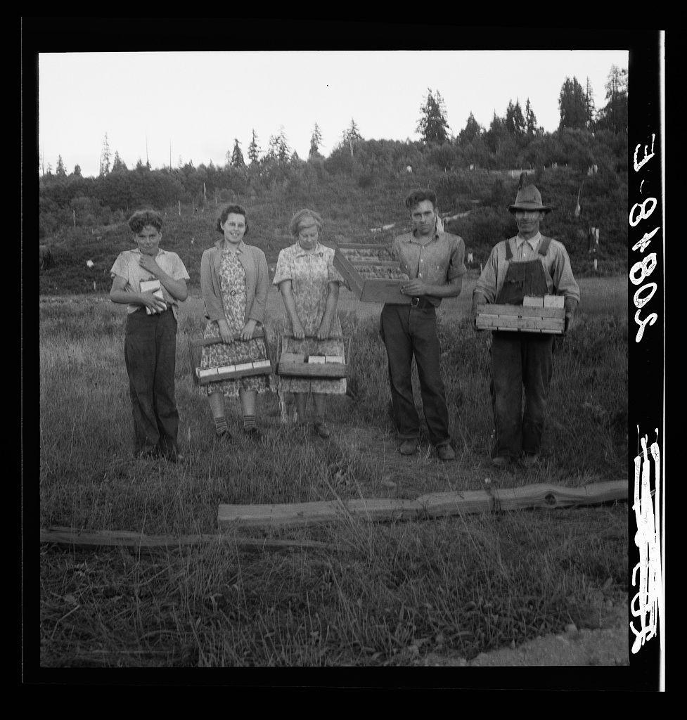 The image is a black and white photograph depicting four individuals standing in an outdoor setting. The backdrop features a natural landscape with grass, trees, and shrubs under a cloudy sky.

From left to right: 
- A young boy wearing suspenders over his shorts holds what appears to be a notebook or book.
- Next stands a woman dressed in patterned clothing holding a bundle of books or papers wrapped together. She has her hands clasped behind her back and is smiling at the camera.
- Another woman, also with a smile, wears a floral dress and sandals; she too is clutching what seems to be several bundles similar to those held by the first woman.

On their right:
- An older man wearing overalls holds a box containing multiple items. He has on boots, gloves, and a hat that appears to have goggles or some sort of eye protection.
The ground they stand upon looks grassy with patches of dirt and what seems like logs laid horizontally across it. The individuals are standing in such a manner as if posed for the photo.

There's handwritten text at both top corners reading "5-8/10". A more elaborate script appears on the right side, which might be part of an inscription or additional context to the photograph but is not clearly legible from this angle.