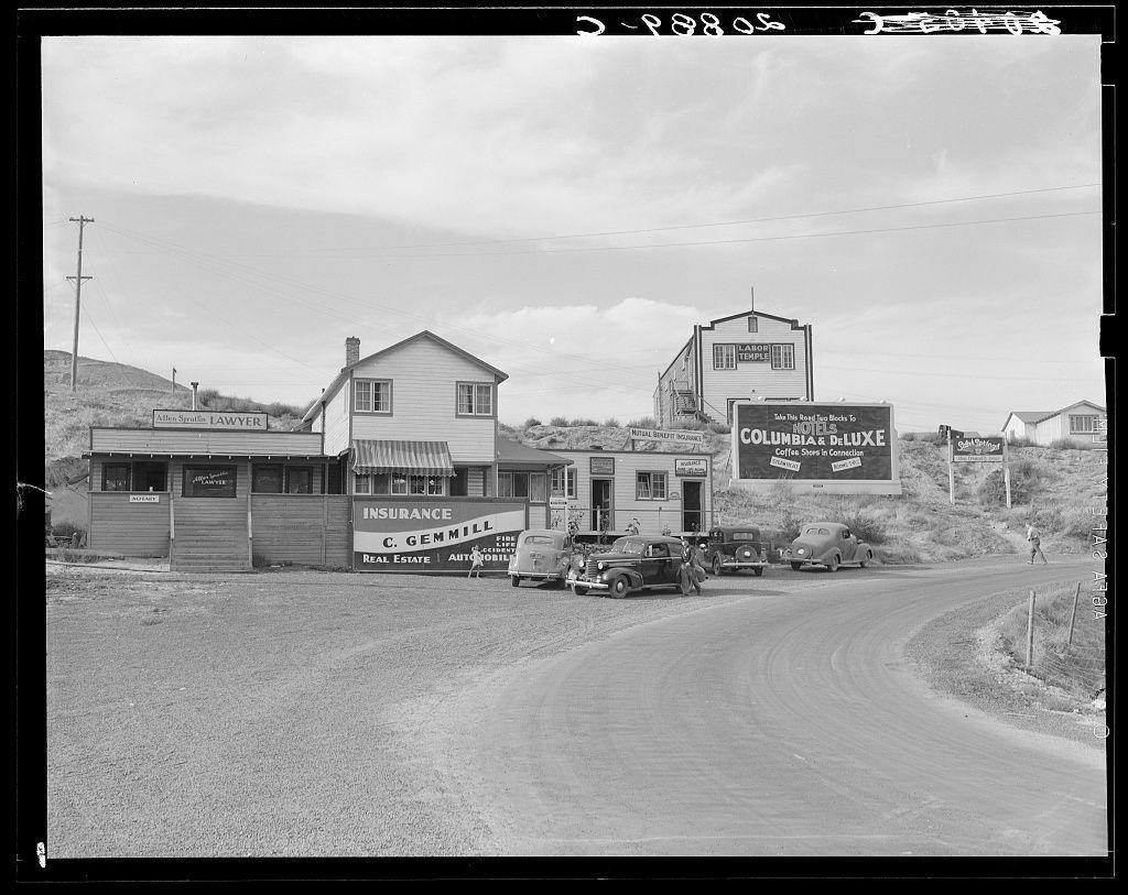 The image depicts a historical black and white scene, likely from the early to mid-20th century. It shows an intersection with several buildings lining one side of the road. There is a mix of commercial establishments such as a lawyer's office labeled "Allen S. Scott," an insurance agency named "C. Gemmill Insurance Co. Real Estate & Auto Mobiles," and another establishment that appears to be related to coffee or hotels, suggesting it might have been a café or hotel with the name "Columbia & Deluxe." A prominent billboard in the background announces promotions for Columbia & Deluxe.

Vehicles from an earlier era are parked along the side of the road. The architecture is simple and functional, typical of small-town commercial buildings during that time period. There's also signage indicating services like real estate, insurance, legal assistance, and what seems to be a reference to coffee or hospitality in connection with "boom construction town," suggesting economic activity tied to infrastructure development.

A person can be seen walking on the road beside one of the parked cars, which is moving away from the viewpoint. The setting appears rural due to open fields visible beyond the buildings and hills further back under an overcast sky. This photograph could potentially relate to Coulee City in Grant County within Washington state as indicated by additional information [...]