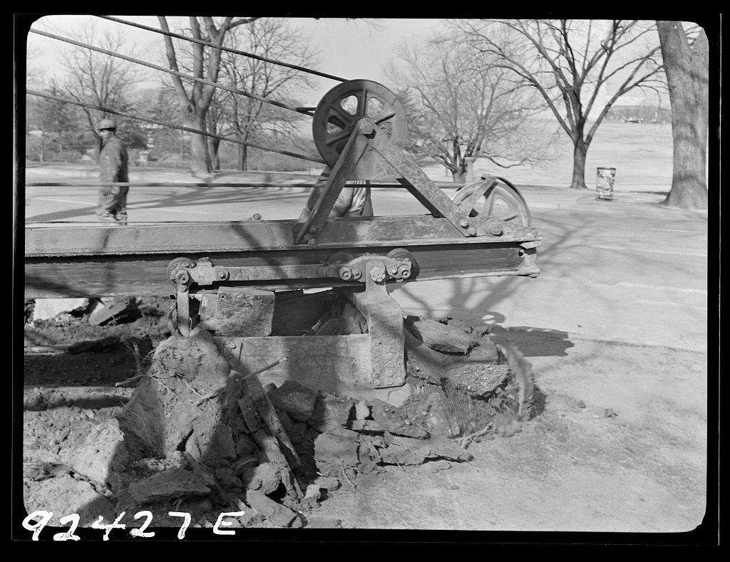 The image is a black-and-white photograph depicting an outdoor construction scene. The focus of the photo appears to be on agricultural machinery, specifically a plow or earth-moving equipment with large metal wheels and gears mounted on what looks like wooden beams. This piece of equipment is situated over a dug-up area with exposed soil and rocks.

In the background, there are leafless trees indicating it could be late fall or winter season. A person in work attire can be seen walking away from the machinery towards the left side of the frame, suggesting labor activity related to construction or maintenance.

The photograph has a caption at the bottom right corner that reads "92437 E," which may refer to an inventory number for archival purposes. The overall setting appears to be in an urban park-like area with open space and pathways visible further back.

Additional information about this image can be found on a website mentioned within the source, suggesting it might have been part of a larger collection or publication related to Washington D.C., specifically concerning earth-clearing and paving work for Independence Avenue.