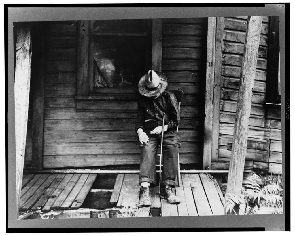 A black and white photograph depicts an elderly individual seated on a porch with one leg raised, resting against the wooden structure. The person is dressed in dark clothing, including jeans, boots, gloves, and a wide-brimmed hat, suggestive of practical or labor-intensive attire. They are holding what appears to be a cane or stick close to their body.
The setting seems rustic, likely from an earlier era based on architectural features such as wooden siding, the simple construction style, and weathered materials indicating age and exposure to elements. The porch is not well-maintained; one plank of wood lies askew beneath where they are seated, suggesting neglect or hardship.

In the background through a partially open door, another figure can be seen in dim light, adding an element of mystery but providing no additional context on their relationship with the elderly person. This image captures a moment frozen in time, reflecting themes of age, solitude, and perhaps economic distress. It evokes feelings associated with rural or impoverished life during a specific historical period.

The photograph is credited to Dorothea Lange (6495), as indicated by its source URL link at the bottom right corner. The mention "Old age" suggests that this image may be part of an exhibition, series, or collection exploring themes around aging in society's context from Washington, Pennsylva [...]