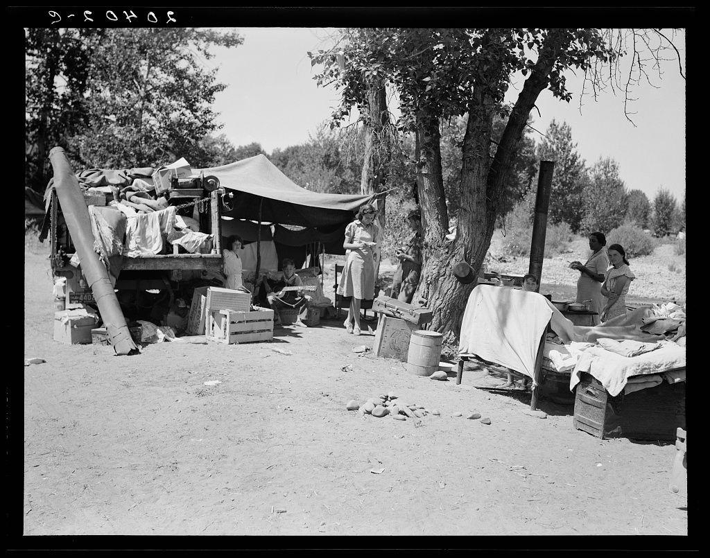 This black and white photograph depicts a makeshift outdoor living area, suggesting an environment of hardship or displacement. The setting includes several people seated around what appears to be temporary shelters made from various materials like blankets, tarps, and fabric. There is visible clutter such as clothing items spread out on the ground, crates filled with unidentified objects, rocks scattered across the dirt-covered floor, a bed draped in white sheets, and a stove or some form of cooking equipment.

The background features sparse vegetation and trees that indicate an arid environment. A vehicle laden with supplies stands prominently to one side, reinforcing the notion of mobility within this encampment setup. The people are engaged in different activities: sitting on wooden benches, standing under the shade provided by a makeshift canopy, or handling personal items.

The overall impression is one of resilience and adaptation amid challenging conditions, as indicated by the caption which references "a family with nine children who have been on the road for three years." This image likely captures a moment in their journey, reflecting historical themes such as migration, displacement, hardship, and survival during times when families faced significant displacement.