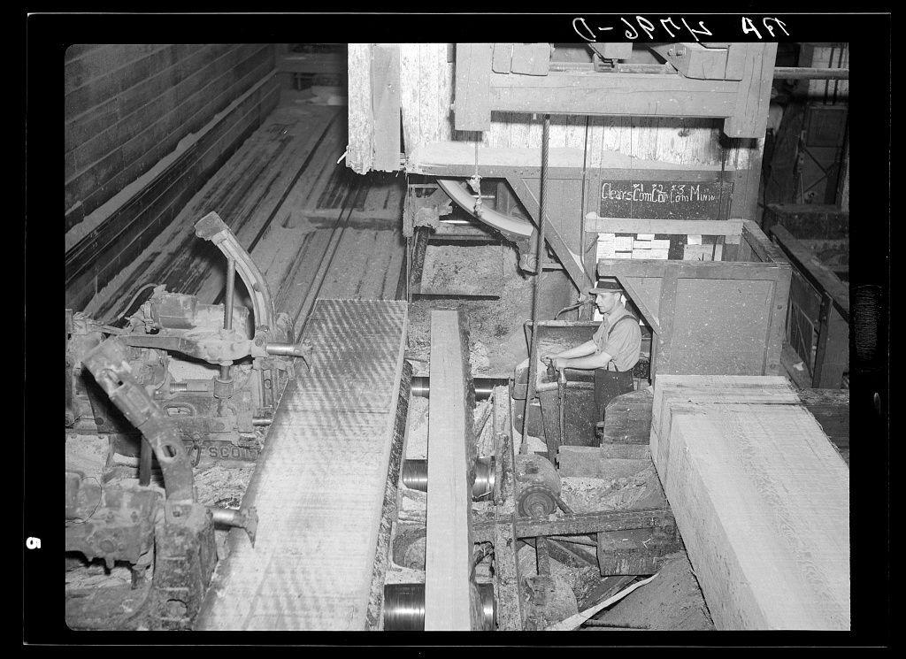 The image depicts a black and white photograph of an industrial setting, likely within the early to mid-20th century. A worker is seated at a large mechanical saw or planer machine, which appears to be processing timber logs into planks or boards. The machinery features various metal components such as gears, levers, rollers, and belts that suggest it's part of an automated milling process.

The environment includes wooden structures in the background, possibly walls made from cut lumber themselves, contributing to a rustic industrial ambiance. There are large rectangular slabs on the ground near the machine where some pieces may be placed after processing or awaiting further treatment.

Notable features include signs with text that read "Clear Cut Longview" and an unclear date marking at the top of the image ("4-SPDR 1937"), hinting at a location, event, or company name associated with this setting. The photograph seems to capture the laborious work involved in milling timber.

The overall scene reflects industrial activity from that era, highlighting manual operation within what could be considered an early factory system focused on lumber processing and possibly paper production given the nature of the machinery used for planing wood into thin sheets or boards suitable for pulp manufacturing.