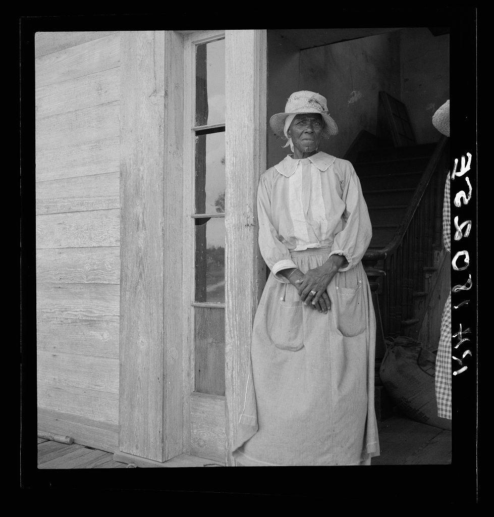 The photograph captures a middle-aged woman standing by an open door on the porch of what appears to be a wooden house. She is dressed in period clothing, indicative of late 19th or early 20th-century fashion: she wears a white blouse with puffed sleeves and a high-necked collar under a wide-brimmed hat adorned with ribbon details. Her long skirt extends down past her ankles and has pockets on either side, suggesting practical use for carrying items.

Her posture is upright but relaxed; she stands sideways to the camera, hands clasped in front of her body, which conveys a sense of calmness or contemplation. The setting appears rural with an unadorned wooden structure serving as the backdrop and another person partially visible on the right side, obscured from view.

The black-and-white tone enhances the historical context and emphasizes textures like wood grain and fabric folds. There's no discernible action taking place; it feels candid rather than posed, offering a glimpse into everyday life of that era in Greene County, Georgia as suggested by the caption indicating its location within the United States.