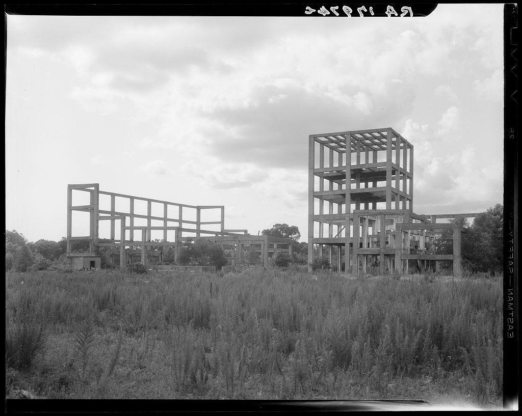 The image depicts a rural landscape featuring skeletal remains of what appears to be an abandoned industrial site. The most prominent structures are large, open-frame frameworks that suggest the remnants of buildings or factories once part of an operational facility. These frames stand in various stages of decay amidst tall grasses and wild vegetation, indicating significant neglect over time.

The sky above is partly cloudy with ample patches of blue showing through, suggesting a day where weather conditions were not overly harsh at the moment the photo was taken. There are no visible signs or indicators that specify what type of industrial plant these remains once belonged to; however, they could possibly be part of an agricultural processing facility.

The overall atmosphere is one of desolation and abandonment, evoking thoughts about the passage of time, economic shifts, and how industries can fall into disuse. The black-and-white nature of the photograph adds a layer of historical context or artistic choice that heightens this sense of nostalgia and loss.