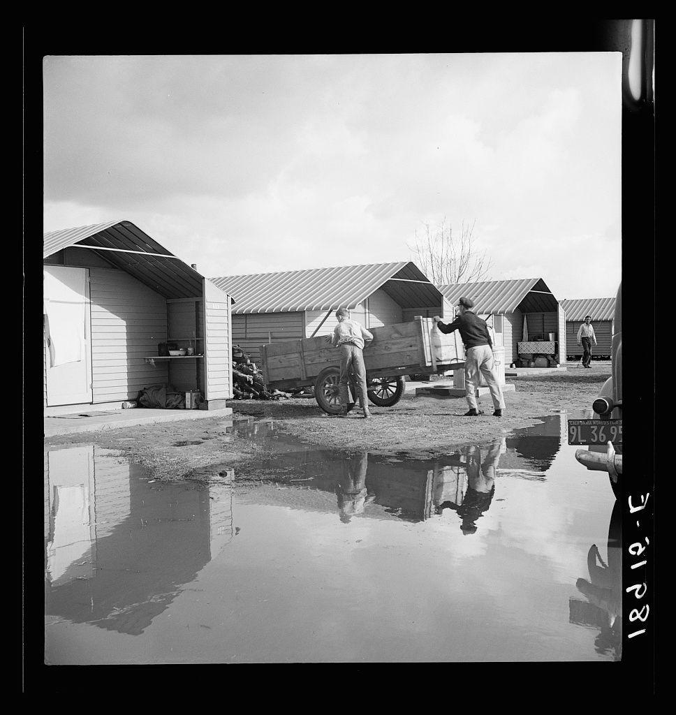 The black and white photograph depicts a scene with prefabricated steel dwellings, known as one step up from a tent. The setting appears to be a temporary or transitional housing area for migrants in Farmersville, California during the Farm Security Administration era. Two individuals are interacting near some small structures and vehicles, reflecting on daily life within this environment. A puddle of water is visible at the forefront, with reflections suggesting an overcast sky above.