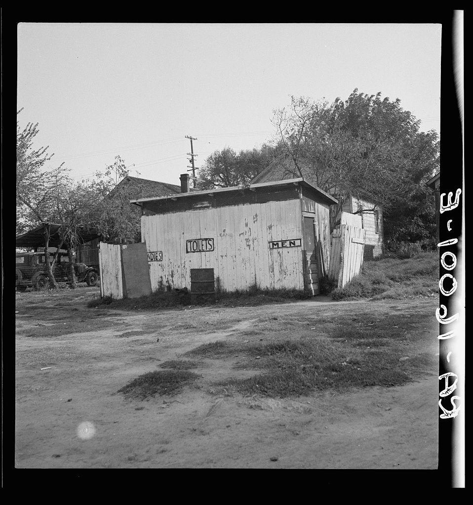 The image depicts a rural outdoor scene with an old wooden structure prominently featuring signs for "TOILETS" and separate sections labeled as "MEN," indicating it's likely used as public restroom facilities. The building shows significant wear, including peeling paint, exposed wood, and general disrepair which suggests abandonment or non-maintenance over time. Surrounding the toilet shed is an expansive dirt area with patches of grass, sparse vegetation, and scattered debris that contribute to a sense of neglect. In the background, there's another wooden structure possibly serving as storage or additional facilities, along with some trees providing minimal greenery in this otherwise barren setting.

A vehicle can be seen parked behind the toilet shed on the left side, reinforcing the notion that this place was once functional but has since fallen into disuse. The photo appears to have been taken during daylight under overcast skies, casting a soft light and minimizing harsh shadows across the scene. Notable is the monochromatic color scheme of black and white which gives it an almost timeless quality.

The presence of such facilities in this setting might suggest that they are located within or near a rural community, perhaps serving workers or residents who may not have access to more modern amenities elsewhere nearby. The historical context indicated by Dorothea Lange [...]