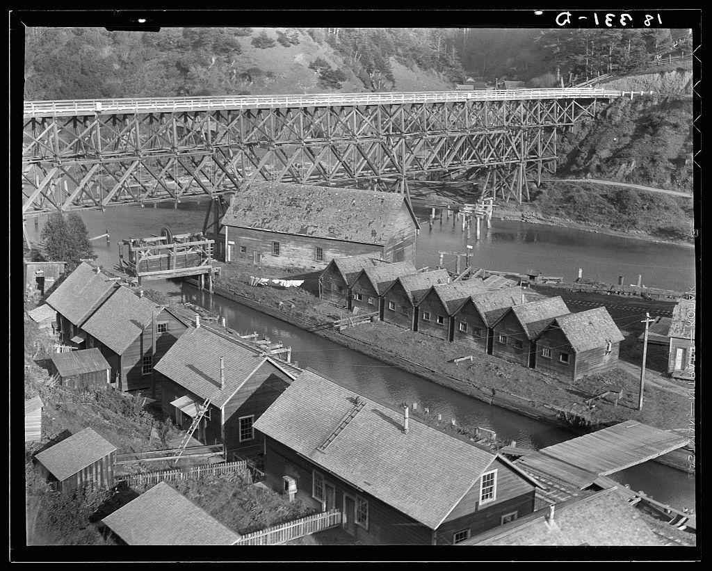 The image depicts an aerial view of a historical site showing the remnants of what appears to be an abandoned lumber town. The focal point is a large wooden bridge with multiple levels, crossing over a river or canal that cuts through the area. Below the bridge and spanning across it are several uniform-looking buildings, possibly former mill workers' housing, characterized by steeply pitched roofs and simple rectangular designs.

The surrounding landscape features various other structures including houses, sheds, and what might be remnants of industrial equipment related to lumber processing. The architecture is utilitarian with a lack of decorative elements, indicative of early 20th-century construction typical for such towns during the height of California's timber industry.

The image appears in black and white, suggesting it was taken or reproduced as an archival photograph from that era. A timestamp "G-168/31" is visible at the top right corner, potentially indicating a cataloging number used by the photographer Dorothea Lange during her documentation of the American West.

In addition to its historical value, this image serves as a visual record for researchers and historians studying California's lumber industry. It offers insight into living conditions in these remote industrial settlements along the north coast of California where timber was once abundant.