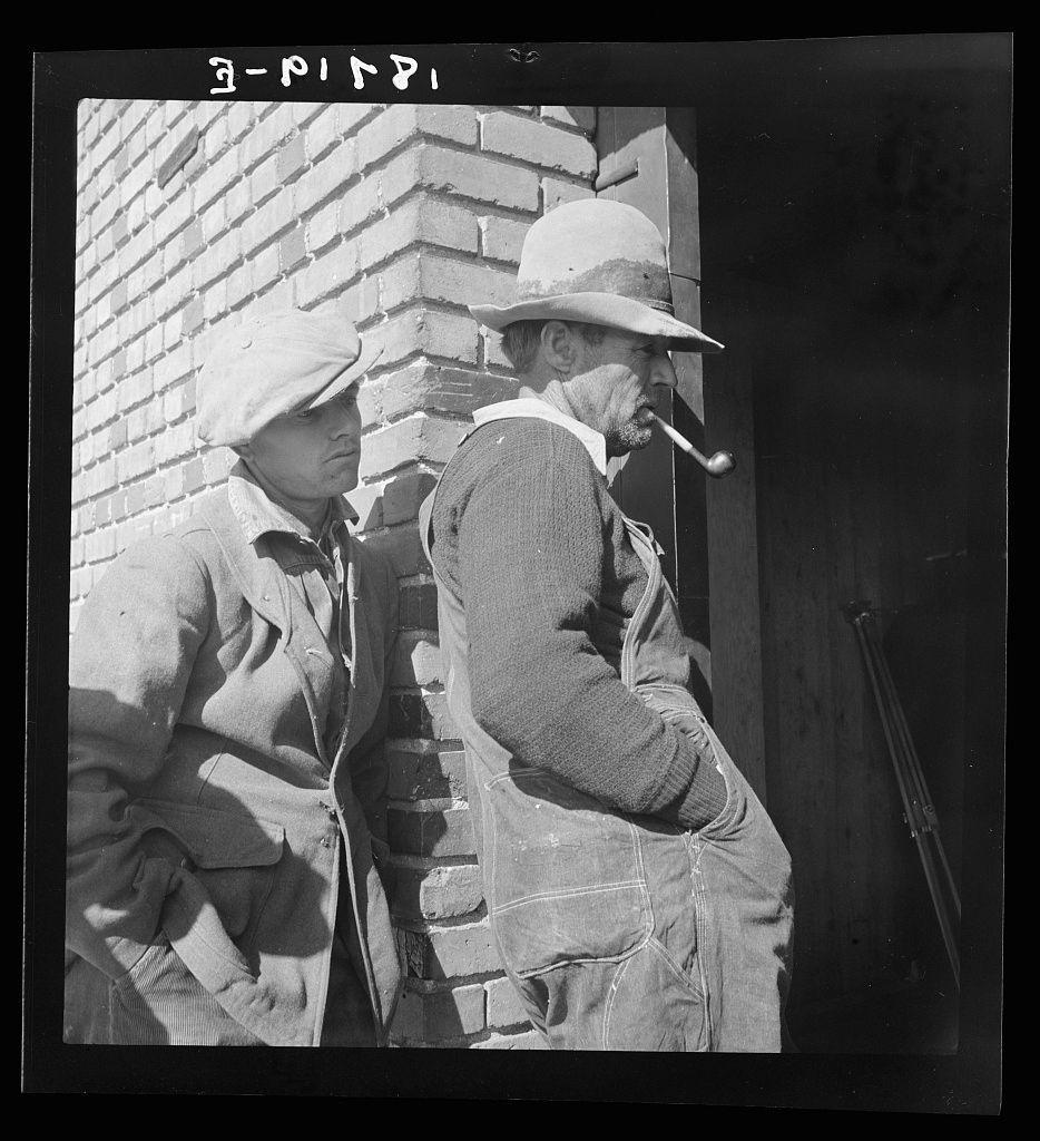 The image is a black and white photograph depicting two individuals in casual attire leaning against a brick structure. The person on the right, likely male based on clothing and posture, appears older with visible facial hair and wears a wide-brimmed hat along with overalls and a sweater vest; he has a pipe clenched between his lips, suggesting an early to mid-20th-century setting given their style of dress. Beside him is another person wearing what seems like work or casual clothes including a jacket and cap. The attire suggests they may be laborers or in some form of manual employment typical for rural workers during the time period indicated by the fashion.

There's no direct indication of race, but both appear to be middle-aged or older men engaged in conversation or contemplation. They stand under sunlight casting sharp shadows on their figures and surroundings, indicative of a sunny day. The presence of such an image suggests it might have been taken during agricultural work hours when laborers would take breaks.

The caption provides context that this photograph is from the Farm Security Administration (FSA), part of its relief program for destitute farm families in Bakersfield, California; indicating these individuals could be beneficiaries or workers associated with such a project. The FSA's mission during mid-century America was to provide aid and support to ru [...]