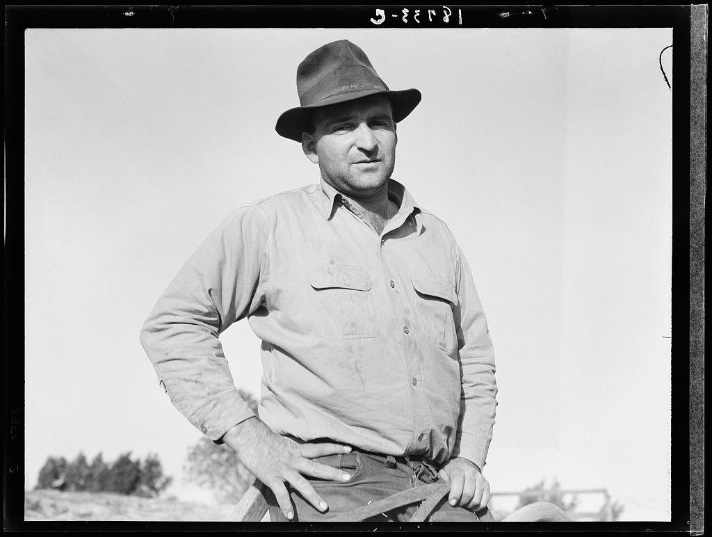 The image depicts a man standing outdoors, hands on his hips and exuding confidence. He is dressed in work attire that includes a long-sleeved shirt with pockets, possibly made of canvas or another sturdy fabric suitable for manual labor. The clothing appears practical and functional rather than fashionable. On top, he wears a wide-brimmed hat which suggests it might be used to protect against the sun—a common accessory among workers engaged in outdoor activities.

The background is minimalistic but gives enough context to infer an agricultural setting; we see sparse vegetation that could signify farmland or rural surroundings under clear skies. The man's posture and expression convey a sense of pride, accomplishment, perhaps resilience as well, which aligns with the narrative provided about him being one among six applicants successful in purchasing his farm through the Bankhead-Jones Act (Tenant Purchase). This act was intended to enable tenant farmers to purchase their land from landlords.

The photograph is monochromatic, typical for its time, hinting at a historical context possibly around early-to-mid 20th century. The absence of color focuses attention on textures and shapes—the rugged fabric of the shirt contrasting against the smooth backdrop of the sky, his polished boots standing out as he leans casually yet firmly onto them.

The presence of trees in the backg [...]