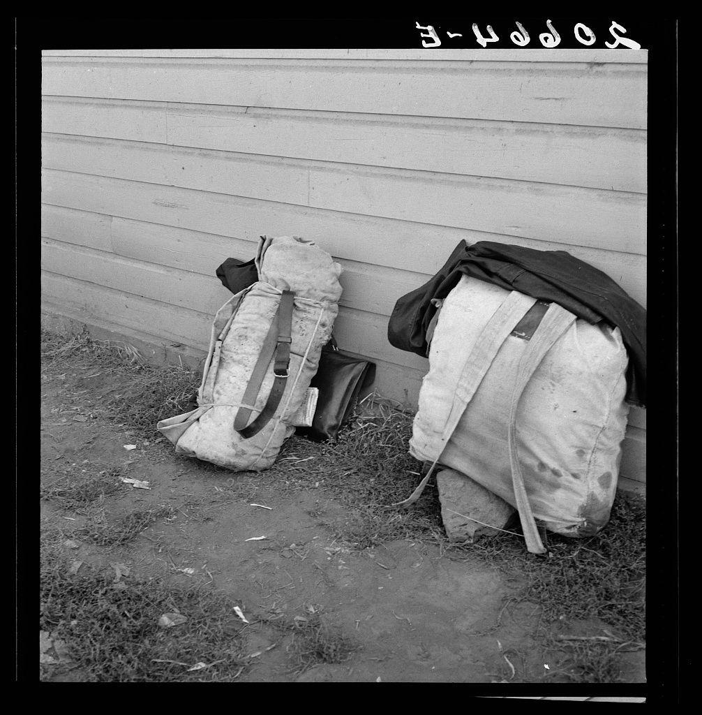 The image is a black and white photograph showing two large backpacks resting against the side of a building. These appear to be military-style backpacks, with one having straps extended outward for support or stability as if someone might have been sitting on it recently.

On top of these backpacks lies what seems like folded clothing: a dark-colored coat is draped over one and possibly trousers or another piece of fabric across the other. The ground beside them looks uneven and covered in patches, suggesting an outdoor setting with some debris such as straw or dry grass scattered around.

The building against which they lean appears to be made of wooden siding, showing signs of weathering but no significant damage. This indicates that it may not have been recently maintained.

There's a stark contrast between the stillness and apparent abandonment suggested by these objects left in this manner outside for an extended period versus what we typically associate with active military or adventure use: readiness to move at any time. The overall atmosphere of the photo is one of neglect, as though someone had to leave hastily without much notice.

The photograph carries a historical feel due to its monochrome format and subject matter - indicative perhaps not only of wartime but also post-war displacement often seen in documentary photography from certain periods (like during  [...]