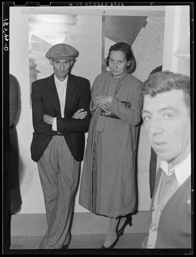 The image is a black and white photograph depicting three individuals, likely from the early to mid-20th century based on their attire. The person in the foreground appears to be an older man with prominent facial features, possibly Dorothea Lange given context clues from provided information about her work during that era.

Behind him stand two other figures; one is a young male wearing glasses and dressed formally in a suit jacket over trousers, standing confidently with arms crossed. Beside him stands another figure who appears to be middle-aged or older, likely female based on clothing style, holding what seems like papers under their arm while donning a long coat.

The background suggests an indoor setting possibly within the context of Dorothea Lange's work for Rural Rehabilitation Division where she documented families applying for farm purchase loans during that time. The individuals in this photograph appear to be part of such documentation process mentioned on a webpage linked provided by source which details about 144 families having applied under Farm Tenancy Act passed by congress in 1937.

No specific actions are depicted, but their composed postures suggest an atmosphere akin to official paperwork or interview.