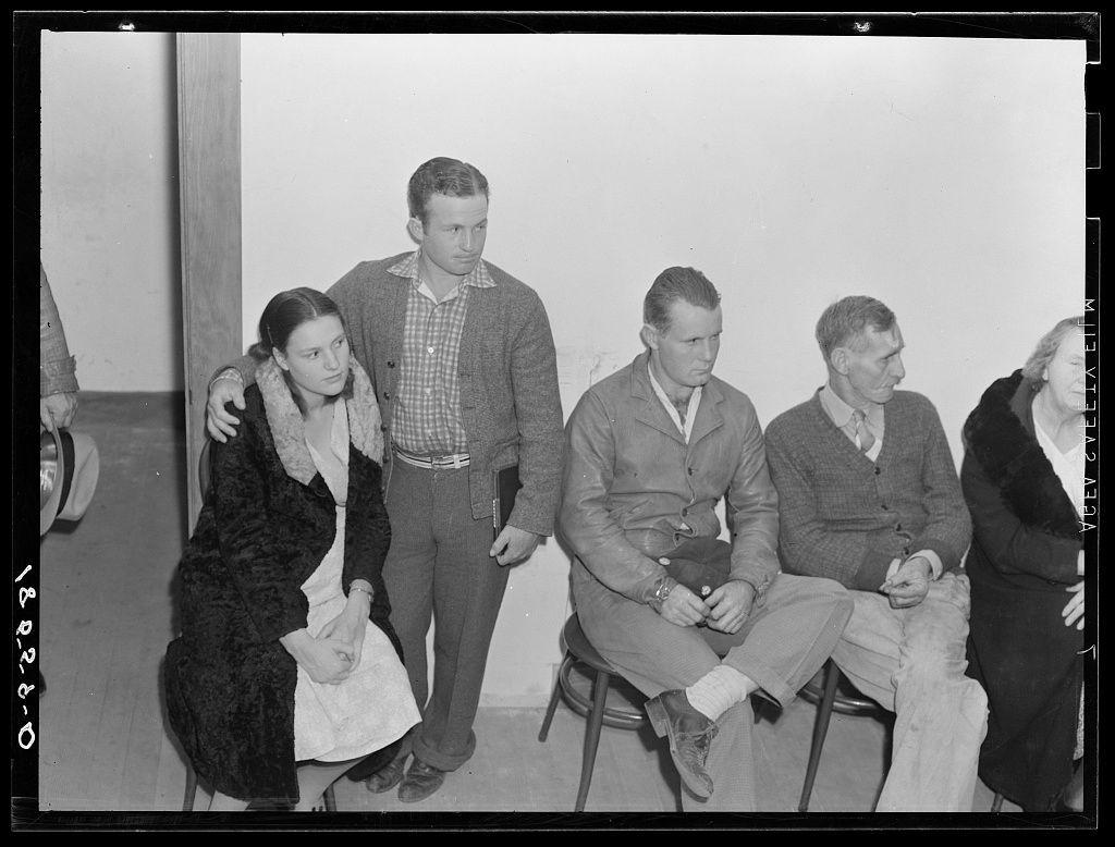 The image is a black and white photograph showing five people sitting in chairs, seemingly waiting for something. They appear to be men and women dressed in modest clothing typical of the mid-20th century. The setting appears to be an indoor room with plain walls.

From left to right: 
1. A woman wearing a dark fur coat over a lighter-colored dress.
2. A man standing behind her, slightly leaning on his hand; he is wearing glasses and casual attire consisting of a sweater, checkered shirt, and jeans. His posture suggests fatigue or weariness.
3. Another seated male with short hair in a darker jacket and trousers, appearing contemplative with hands clasped together.
4. A third man sitting next to the first, dressed similarly; he is also looking down at his shoes with an attentive expression.

5. On the far right edge of the photo: An older woman wearing glasses and a dark coat appears partially out of frame.

The photograph captures a moment that likely relates to agricultural or rural community affairs, given the context provided in the additional information about Night meeting in the Farm Security Administration (FSA) office for farm purchase loans. The image evokes a sense of historical social documentary work focusing on people and their experiences during a specific period related to rural life and economic activities.