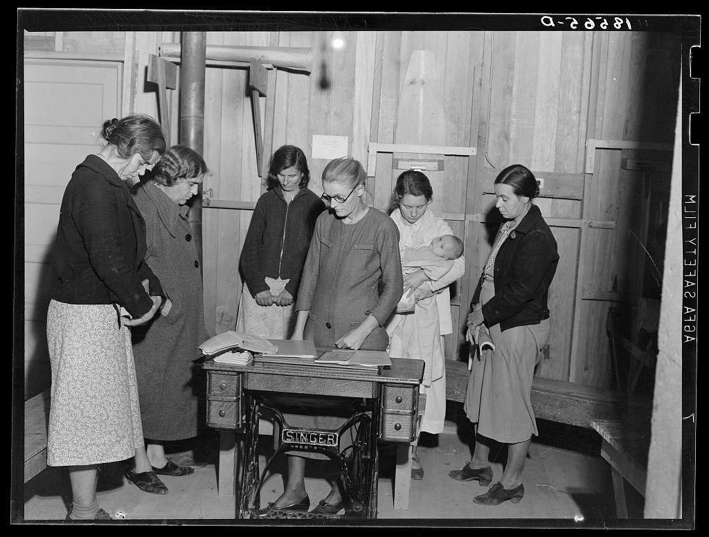 The image is a black and white photograph showing six women gathered around an old-fashioned Singer sewing machine. They appear to be in an indoor setting, possibly within a barn or similar structure given the wooden walls and beams visible in the background.

Five of the women are wearing coats and skirts typical of early mid-20th century fashion, suggesting that this photo may have been taken during that era. One woman is holding an infant wrapped snugly against her chest with a white fabric covering most of its body for warmth or modesty.

The expressions on their faces range from attentive to smiling, indicating they are engaged in conversation and possibly enjoying the meeting. The group seems informal and congenial. There's also a sense that this gathering has some significance; perhaps it is part of a club activity as indicated by one woman wearing an apron with a badge or emblem.

The photo captures a moment from the past, depicting what appears to be a social event among women who are likely mothers given their attire and engagement in activities like sewing. This aligns with historical records indicating that migrant workers' camps often included communal spaces where families could gather for various purposes including socializing and children's care services.

The precise location of this photograph is identified as the Arvin camp, a Farm Security Administrati [...]