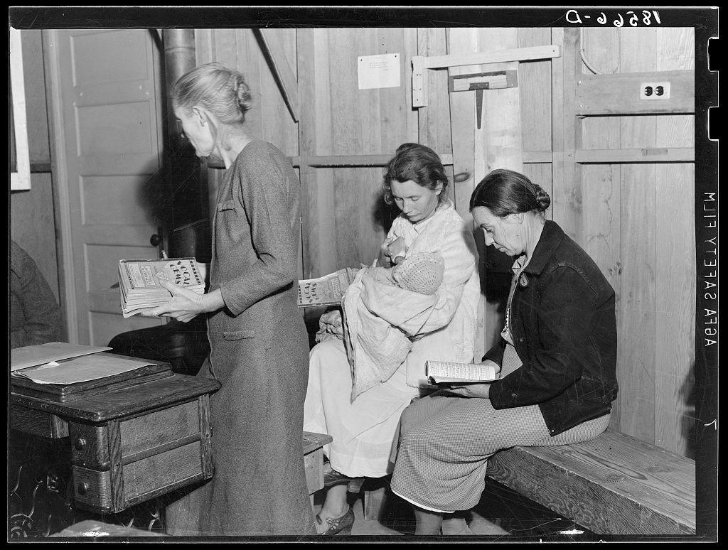 The image captures a moment from the Hymn singing at meeting of Mothers' Club in Arvin migrant camp, California. It is presented as a black and white photograph showcasing three individuals: an older woman standing on the left side holding sheet music with glasses perched atop her head, another elderly woman seated on a wooden bench cradling a newborn baby wrapped in blankets, while yet another female figure sits beside them engrossed in reading from a book. The setting suggests a communal gathering within a modest structure characterized by its simple architecture and utilitarian furniture such as the old-fashioned desk and metal scale mounted on the wall behind the group.