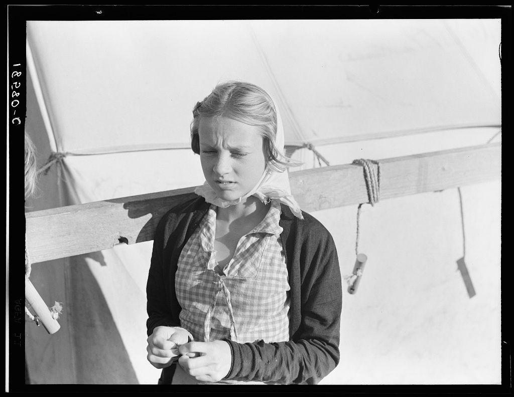 The image is a black and white photograph of a young girl standing in front of what appears to be a tent. The setting suggests that the location may not have modern amenities, hinting at an environment where living conditions are basic or temporary.
In terms of clothing, she wears layers: a dark cardigan over a checked shirt with rolled-up sleeves, suggesting it might be warm enough for short-sleeved shirts but cool outside due to wind. The girl's expression is pensive and somewhat troubled; her eyes gaze downwards as if lost in thought while holding what could possibly be knitting needles or other similar tools.
The surrounding environment lacks detailed features that would suggest a specific context, leaving much open-ended interpretation-wise regarding the story behind this photograph. There are no clear indicators of time such as modern clothing styles or technology present to provide further contextual clues about when exactly it was taken.