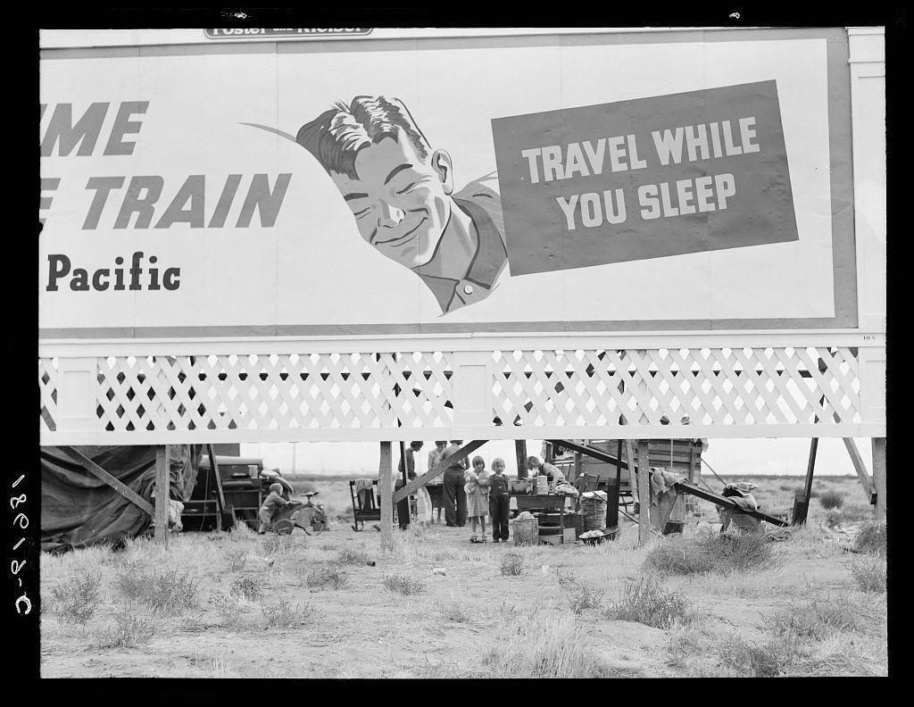 The image displays a black and white photograph of an outdoor billboard advertisement. The ad features bold, capitalized lettering that reads "HOME TRAIN Pacific" with the phrase "TRAVEL WHILE YOU SLEEP" in smaller letters beneath it. An illustration depicts a smiling man's face partially emerging from behind what appears to be the corner of his train sleeper berth window. Below this graphic is more text stating "Pacific." The background shows an outdoor setting where several individuals, including children and adults, are gathered around makeshift structures that resemble tents or temporary shelters made with tarps and other materials. These people seem to be partaking in various activities such as working on construction tasks, conversing, or engaging in daily chores. A bicycle is also visible near the left side of the photo. The setting suggests a rural area, possibly indicating hardship conditions due to displacement or migration.