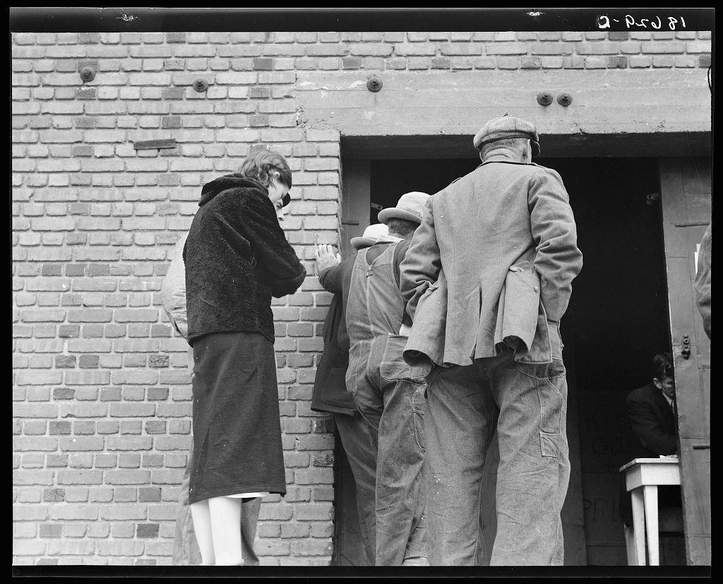 A black and white photograph depicting four individuals standing outside a brick building. The woman on the left, wearing glasses, dark coat, skirt, appears to be conversing with another individual inside through an open door. A man in overalls is partially visible from behind; he seems engaged in conversation or looking at something out of frame. Another man stands beside him, facing away and dressed in a suit jacket, trousers, cap, suggesting casual yet somewhat formal attire for the era depicted. The scene suggests social interaction during what might be an application process within a government-operated food grant distribution depot operated by the Farm Security Administration (FSA), specifically located in Kern County, California as documented on archive.org with accompanying text describing it as part of a series showcasing destitute farm labor families seeking aid from FSA. The photograph carries historic significance indicating a time likely during or around WWII era based on clothing styles and context provided by the archival information associated with this image.