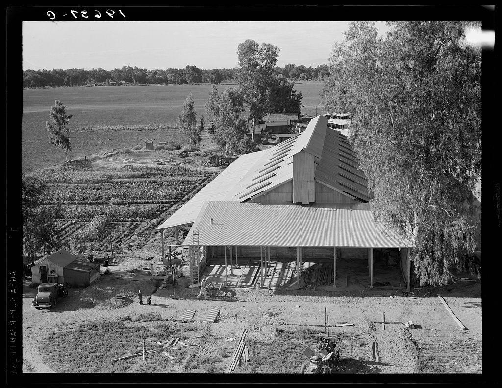 The image depicts a rural landscape featuring an old-fashioned, weathered barn with a prominent roof structure. It is situated in what appears to be the mid-20th century based on architectural styles and black-and-white photography quality. There's visible construction or renovation work around the building. Surrounding this main feature are open fields indicative of agricultural land use, with various farming tools scattered across the grounds. The environment suggests a dry climate due to sparse tree cover and barren landscape. In the foreground, there is an old-fashioned vehicle that adds historical context. Overall, it captures rural life possibly in California during a time when barns were central to farm operations before modern agricultural machinery changed practices significantly.