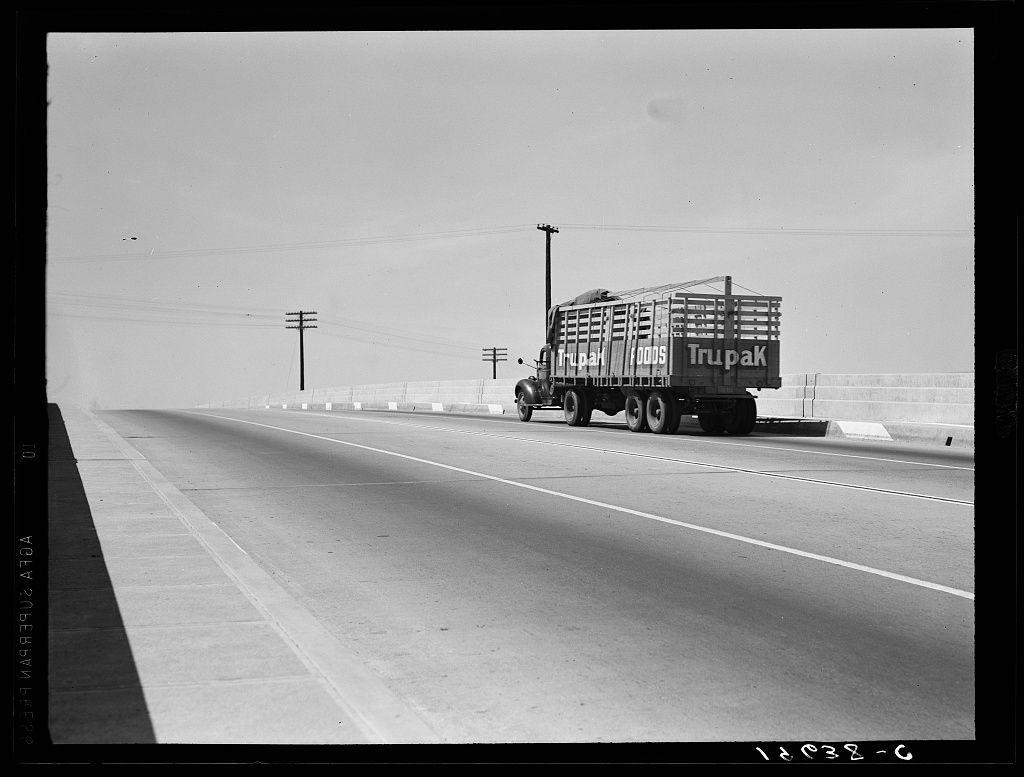 The image is a black and white photograph depicting an overpass on U.S. 99 in California, located between Tulare and Fresno. It shows a large truck driving along the highway with its cargo secured within a fenced enclosure labeled "Trupak." The vehicle's design indicates that it was likely taken several decades ago, reflecting mid-20th-century transportation aesthetics.

The road appears to be an interstate or major thoroughfare, given the wide lanes and concrete barriers. Telephone poles are visible along the sides of the highway, contributing to a sense of depth in the composition. The sky is clear with no clouds present, suggesting fair weather conditions at the time of photography.