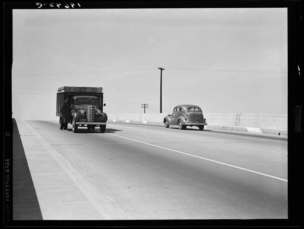 The image captures a black and white scene of two vintage vehicles traveling on what appears to be an overpass, with clear skies above. On the left side is a large truck hauling goods in its enclosed cargo area; it features pronounced fenders at each wheel well, suggesting robustness for heavy-duty transport. The right vehicle seems to be a passenger car from roughly the same era as the truck, distinguished by rounded bodywork and classic automotive design elements like prominent headlights and an open grill.

In terms of context provided in the caption "Untitled photo," this image is likely connected with California's transportation infrastructure during mid-20th century. The overpass on U.S. Route 99 hints at a significant route connecting Central Valley cities Tulare and Fresno, indicating that these vehicles are part of daily life or commerce along major interstate highways. This particular photograph could be emblematic of the broader social history depicted in Dorothea Lange's work during the Great Depression era when she documented various facets of American society.

Lange is known for her evocative and poignant imagery capturing moments from everyday existence, particularly those marked by hardship or resilience against such challenges—themes that resonate with this snapshot. The absence of modern traffic signals, road signage, or contemporary infrastructure elem [...]