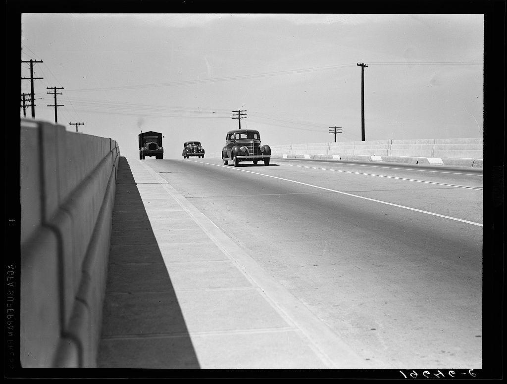 A black and white photo shows three vintage cars driving down a highway. The road is flanked on both sides by concrete barriers, with telephone poles visible in the distance. There are no people or animals present in this image. This photograph appears to be part of Dorothea Lange's work captured between Tulare and Fresno along U.S. 99 in California as indicated in a caption at the bottom right-hand corner of the photo which reads "Lache-6."